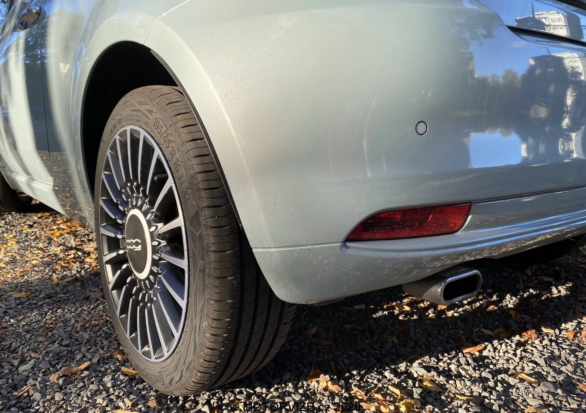 Közeli kép egy világos színű 2015-ös Fiat 500 hátsó negyedéről, alacsony szögből készült fotó. A többküllős alufelnire és a látható mintázatú gumira fókuszál, ami egy sportos modellre utal. A hátsó sárvédő elegáns íve és a fémes karosszéria matt tükröződése látható. Látható egy rész a LED hátsólámpából és a krómozott kipufogóvégből. A parkolószenzor és a hátsó ködlámpa fejlett biztonsági funkciókra utal.