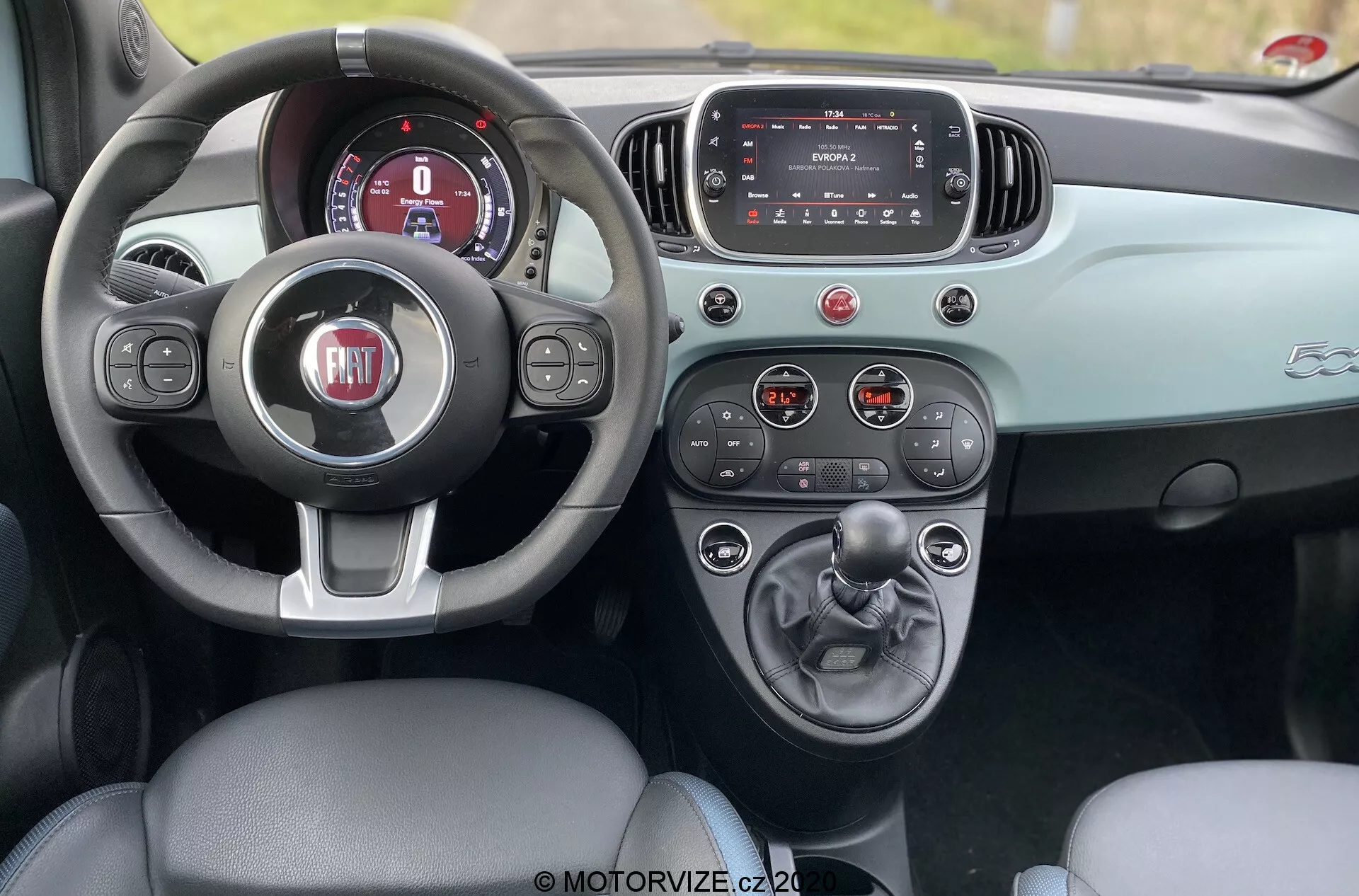 Egy 2015-ös Fiat 500 belső felvétele a hátsó üléssorból, amely bemutatja az első pilótafülkét bőr sofőrüléssel, háromküllős kormánykerékkel a FIAT logóval, digitális műszerfallal, infotainment érintőképernyővel és középkonzollal kézi sebességváltóval és pohártartókkal. A műszerfal és az ajtó részletei kontrasztos kéttónusú színsémát mutatnak, amely fokozza az esztétikai designt. A kép nappal készült, kiemelve az autó kifinomult belső kialakítását és funkcionalitását.