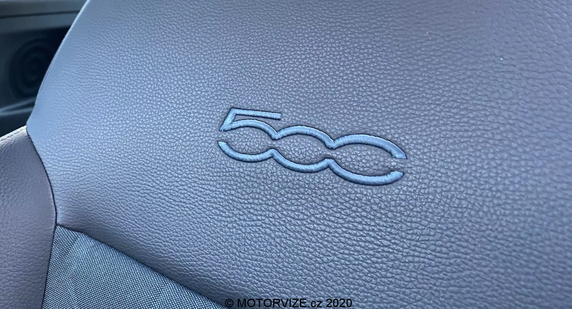 Közelkép a Fiat 500 2015-ös modell belső terének műszerfalán stílusosan domborított '500' logóról. A bőrhatású textúrát és a műszerfal panel precíz varrását a megvilágítás és a szűk mélységélesség emeli ki, hangsúlyozva a jármű belső kialakításának magas minőségű kivitelezését.