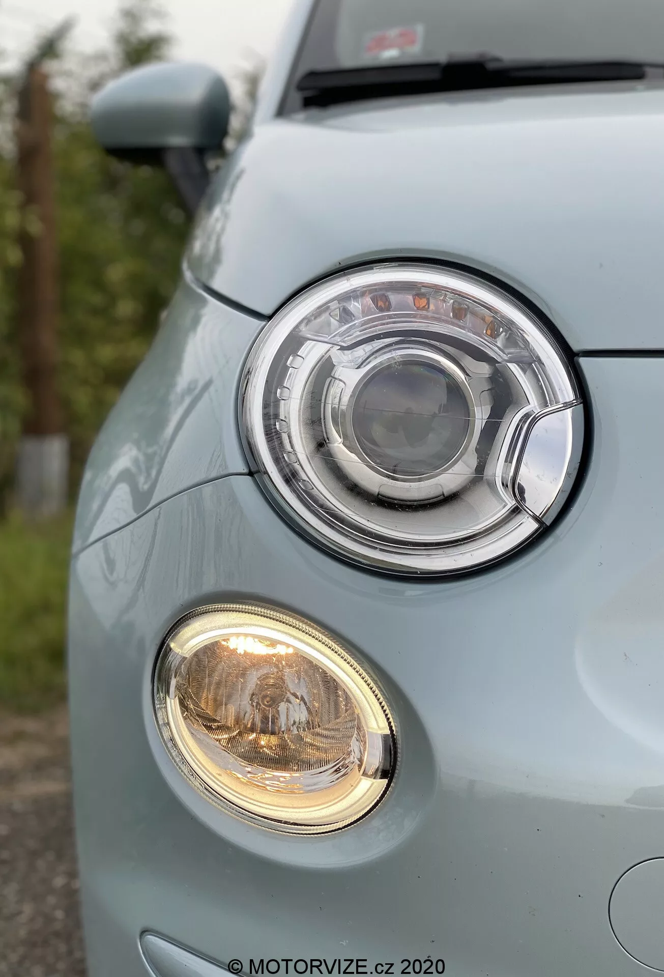 Egy világoskék Fiat 500 2015 elülső bal sarkának közelképe, amely kiemeli a halogén projektoros fényszórót egy körkörös kialakításon belül, amelyet egy króm gyűrű vesz körül. Alatta egy kisebb ködlámpa meleg, sárgás fényt áraszt. Az alacsony szögből készült kép kiemeli a sima és lekerekített első lökhárítót egy jellegzetes rácsozattal és rendszámtábla-területtel, hangsúlyozva a jármű jól karbantartott, városi orientációjú kialakítását.