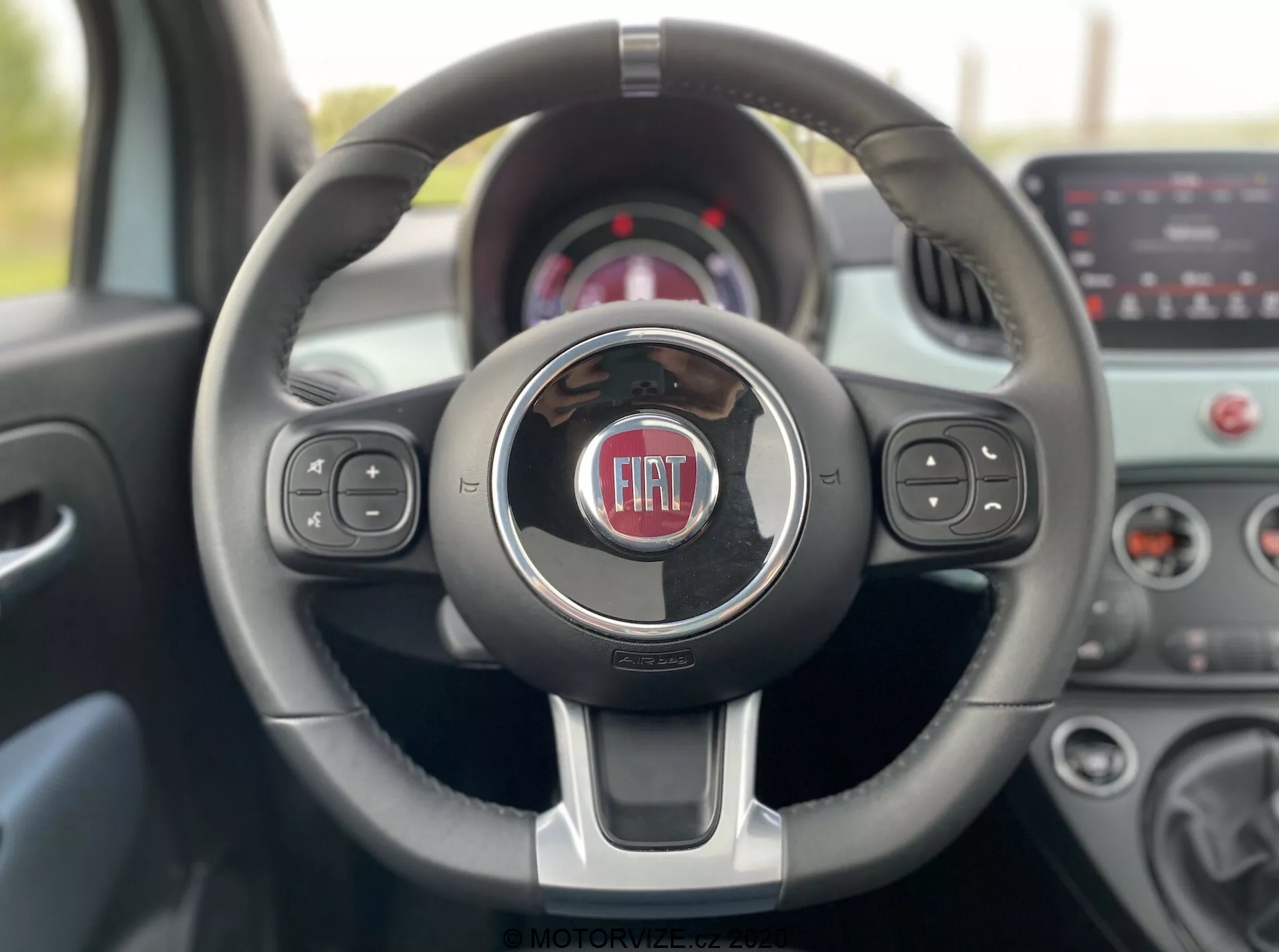 A 2015-ös Fiat 500 beltéri nézete a vezetőülésből, amely egy modern, háromküllős kormánykereket mutat króm díszítésekkel és a Fiat logóval középen. A kormányon médiavezérlők, hangvezérlés és sebességtartó automatika gombjai találhatók. Az elhomályosított háttér műszerfalat és érintőképernyős infotainment rendszert tartalmaz, kiemelve a jármű kortárs technológiáját és designját.