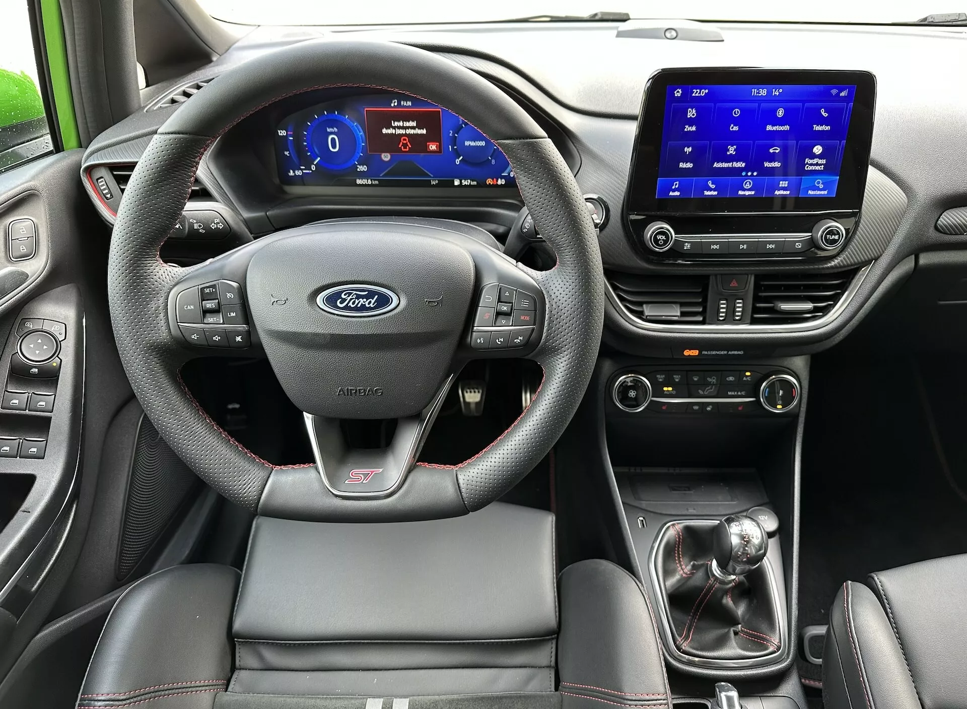 A 2022-es Ford Fiesta belsejének nézete a hátsó ülésről, kiemelve a Ford logóval és ST jelzéssel ellátott kormánykeréket, digitális műszerfalon keresztül, érintőképernyős infotainment rendszer, kézi sebességváltó, és a kontrasztos varrással ellátott sportos vezetőülést.