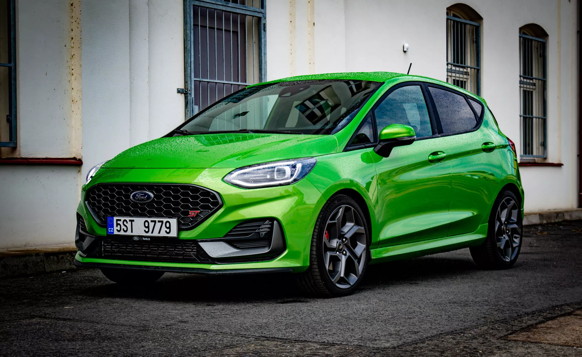 2022-es Ford Fiesta élénk zöldben, amely sportos tervezését mutatja be alacsony első háromnegyedes nézetből, kiemelve az első lökhárítót, hűtőrácsot, fényszórókat, motorháztetőt, sárvédőt, kerekeket és a szélvédő visszatükröződését városi utcán az eső után
