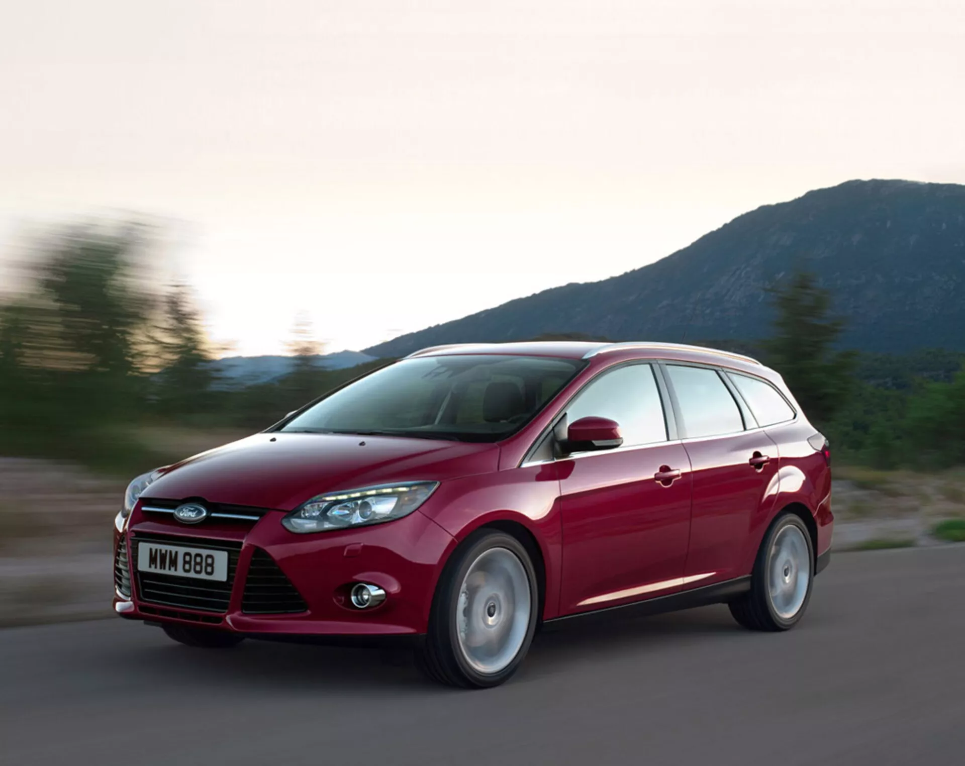 2011-es piros Ford Focus kombi úton, dinamikus designnal, kiemelkedő első ráccsal és Ford logóval, hátrafelé hajló fényszórókkal, formált motorháztető vonalakkal és ötvözet kerekekkel, esti fényviszonyok között