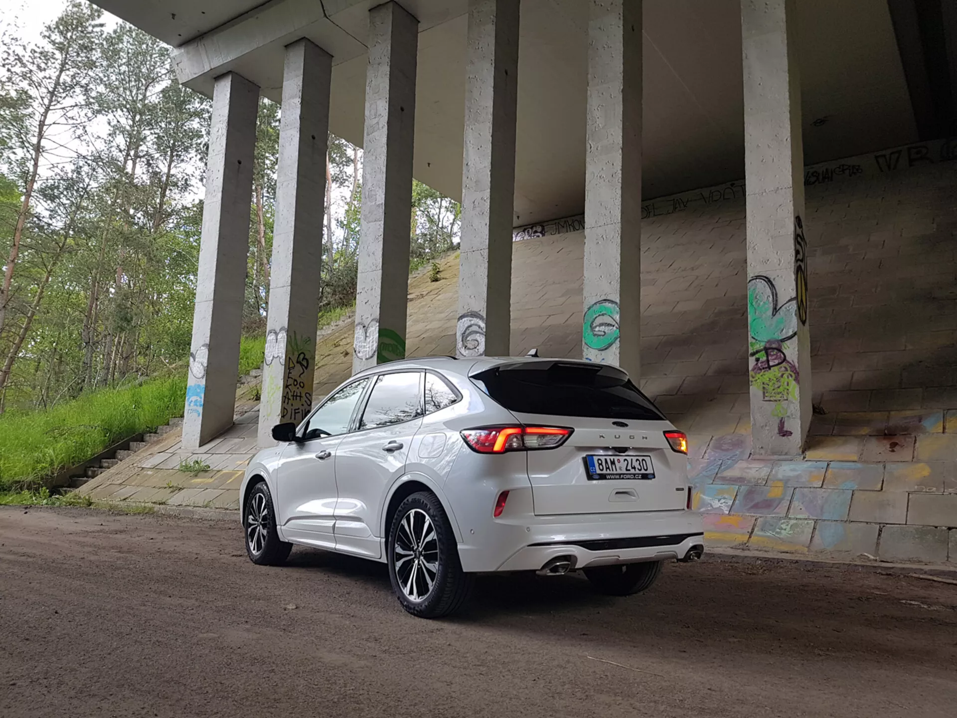 2019-es Ford Kuga crossover SUV parkol egy felüljáró alatt, bemutatva modern tervezését és technológiai jellemzőit, mint az LED hátsó lámpák, kettős kipufogó és tolatókamera, városi graffitik és betonoszlopok között