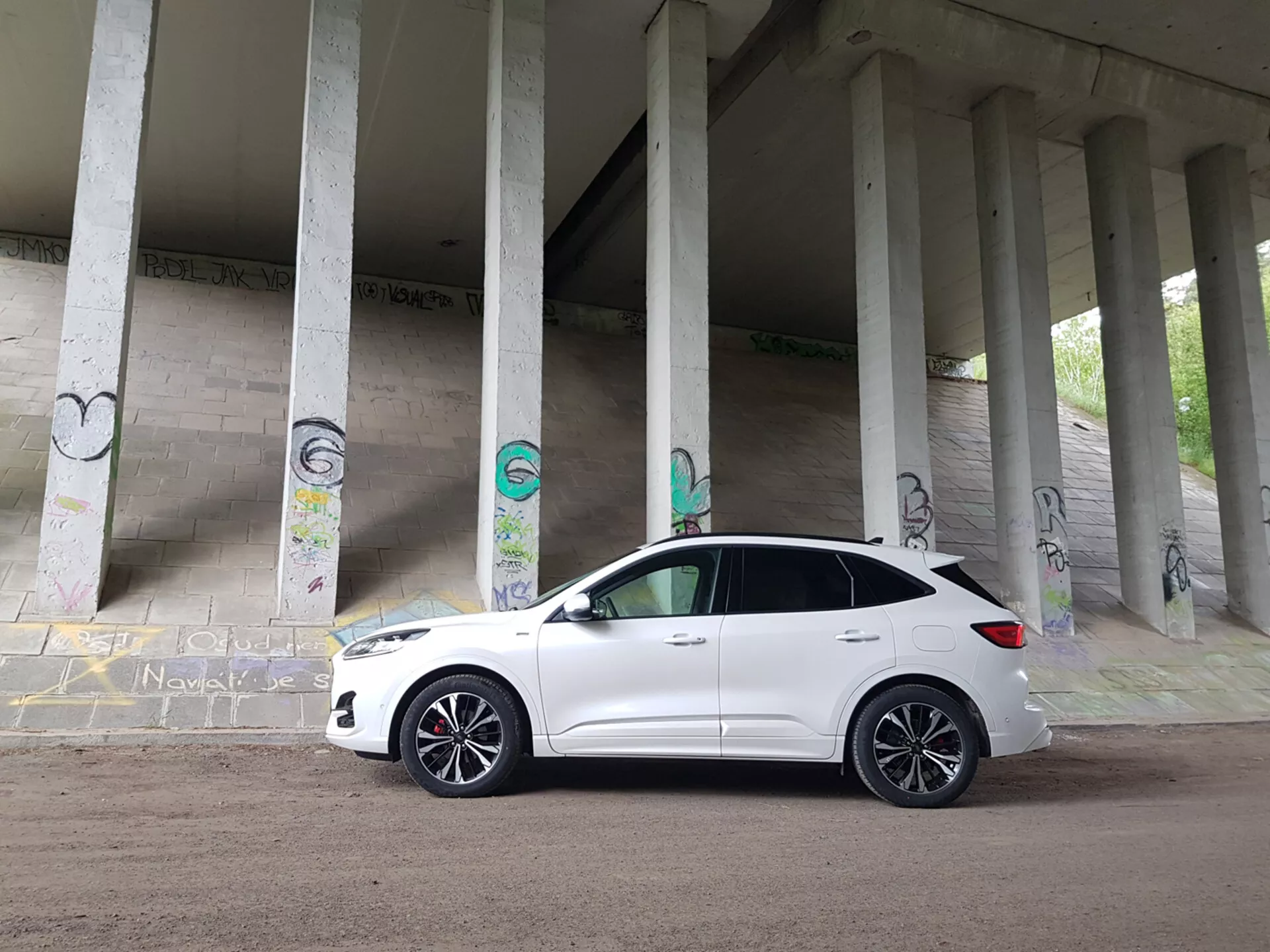 2019-es fehér Ford Kuga kompakt SUV, graffiti borította betonoszlopok alatt parkolva, bemutatva az első negyed profilját, beleértve a hűtőrácsot, a fényszórókat és az ötvözött kerekeket