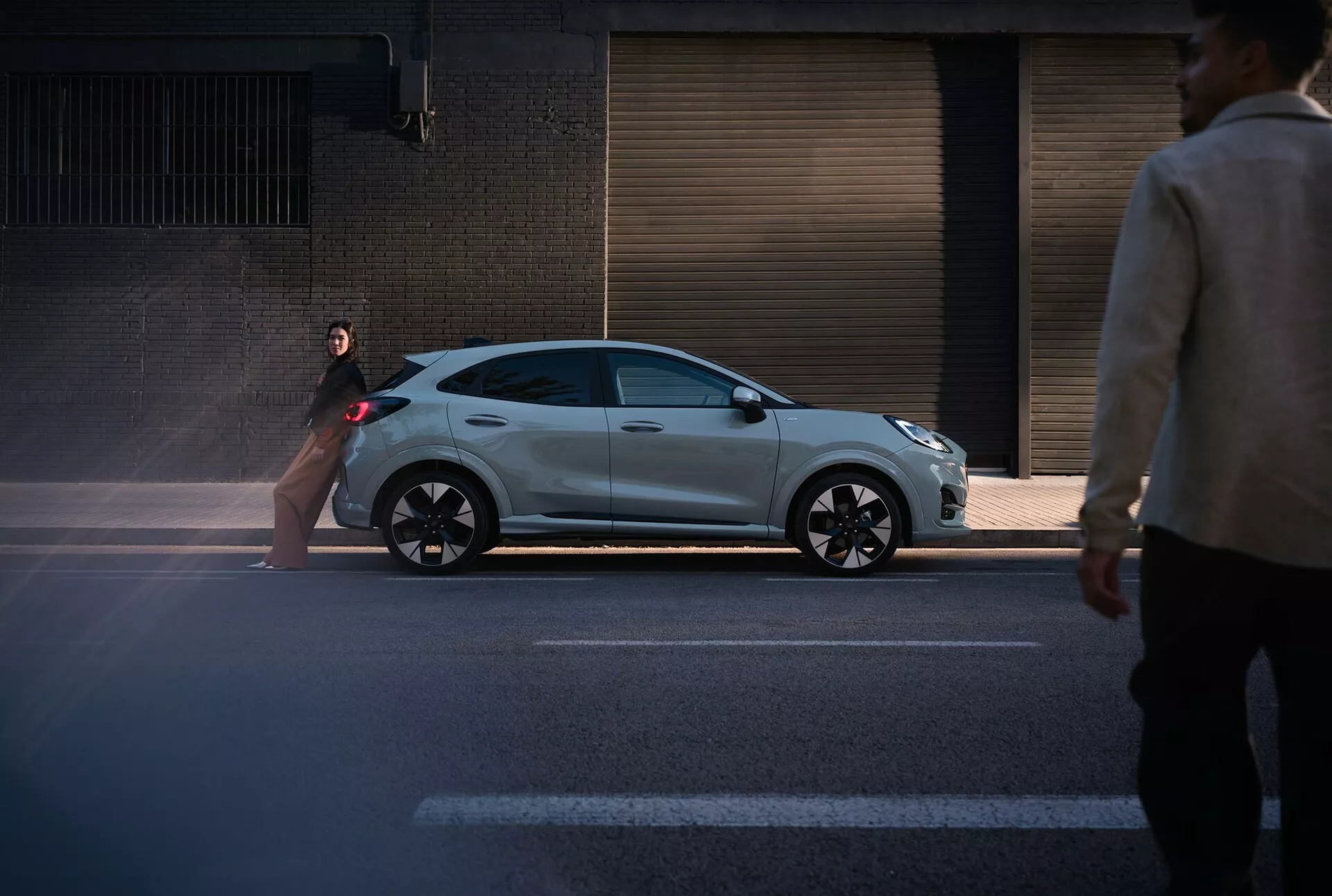 2024-es Ford Puma (II-facelift) ferdehátú metalikus ezüstben, sportos kialakítással, az utca szintjéről készült oldalelnézeti felvétellel, mely bemutatja csinos profilját és modern jellemzőit, mint például a fekete részletmegoldások és többspókás kerekek egy városi háttérrel