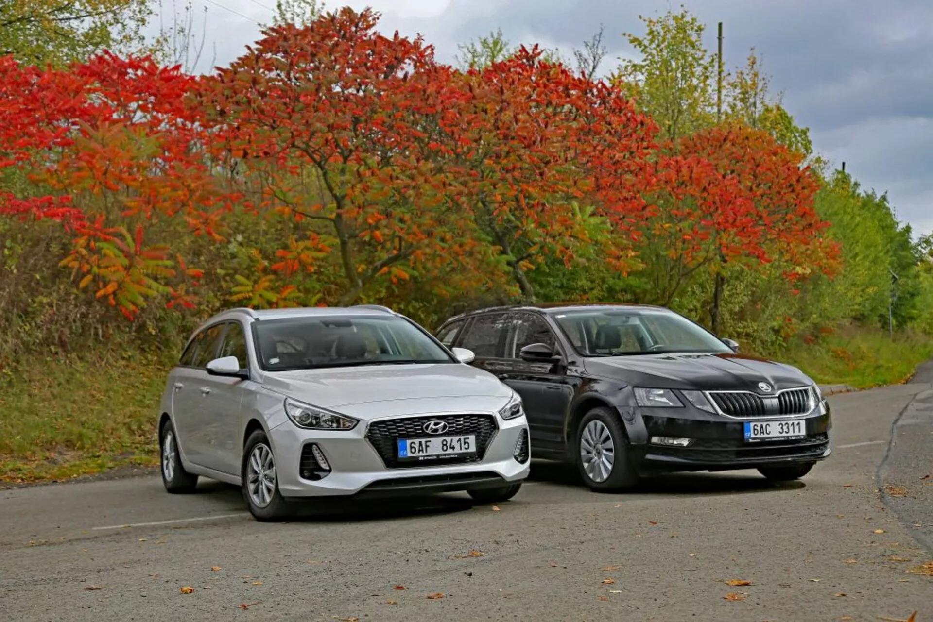 2017-es fehér Hyundai i30 hatchback és sötétszínű szedán egymás mellett egy csendes úton, őszi növényzet által körülvéve, bemutatva a tervezési vonalakat és a modern világítástechnológiát