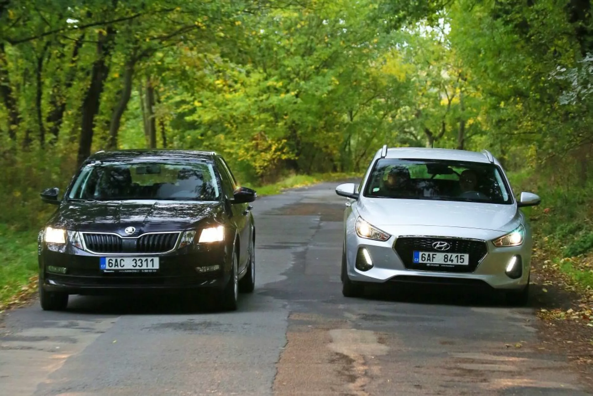 Két autó szemben áll egymással egy fákkal szegélyezett vidéki úton a kora ősz folyamán: egy sötétszínű Skoda téglalap alakú ráccsal és szabványos fényszórókkal balra, és egy világosszínű Hyundai i30-N ívelt elejével, modern projektorszerű fényszórókkal és integrált ködlámpákkal jobbra, bemutatva a hagyományos és modern autótervezési elemek összehasonlítását.