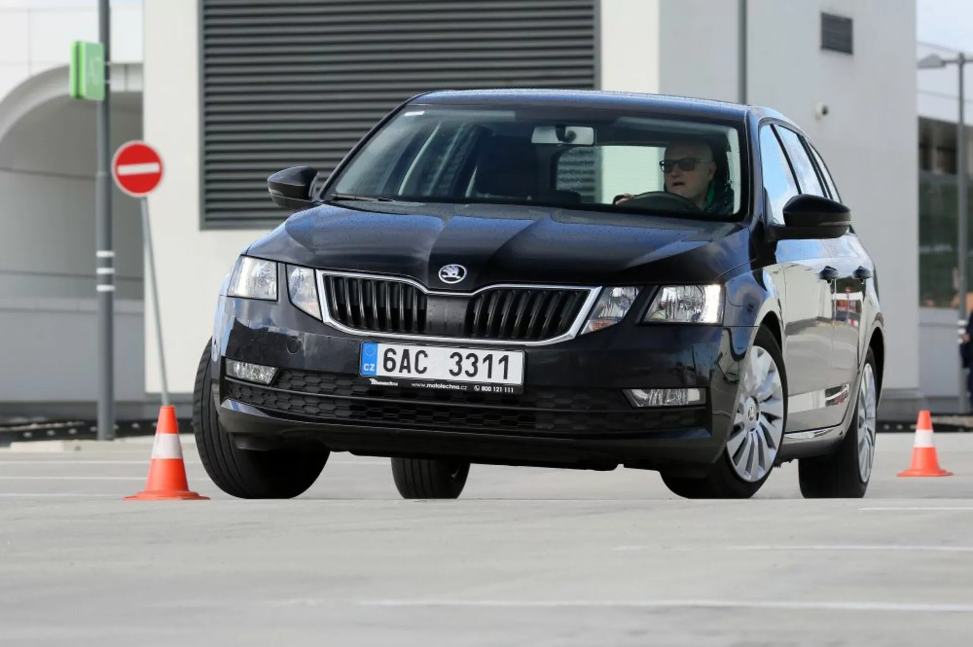 2017-es fekete Hyundai i30 négyajtós szedán, amely narancssárga forgalmi kúpok között manőverezik, bemutatva a jármű kezelhetőségét, az első-háromnegyed nézetből látható a gyártó logója a hűtőrácsnál és a halogén vagy xenon fényszórók LED minták nélkül, hagyományos szélvédőmosó technológia, mozgásban lévő kerékek, amelyek mozgást sugallnak, és nem látható luxus kiegészítők vagy fejlett technológiai jellemzők, egy aszfaltozott tesztterületen