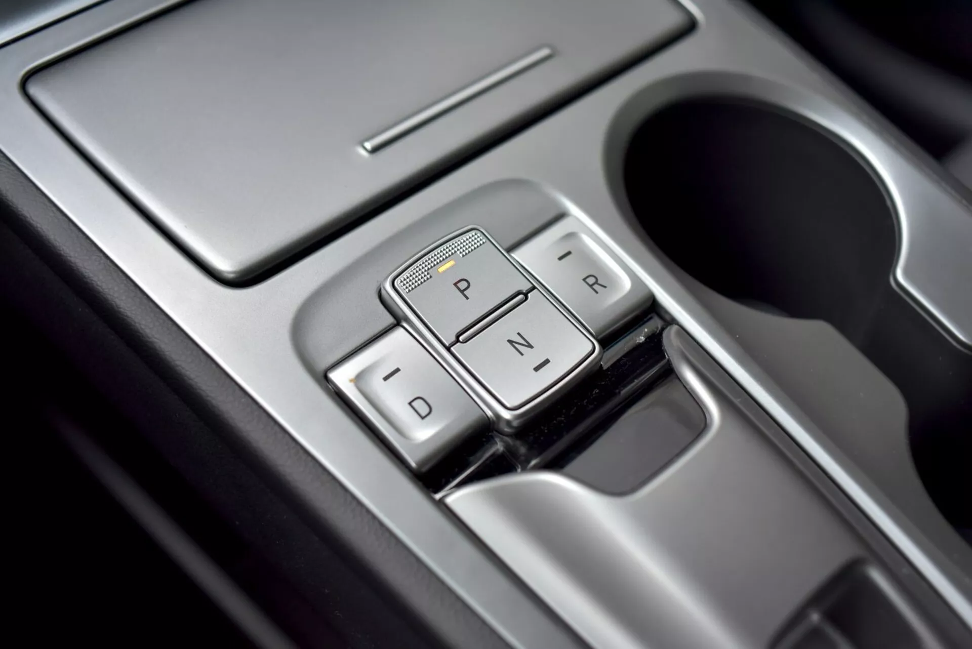 Közeli nézet a Hyundai Kona 2021 modern elektronikus váltókar mechanizmusára a középkonzolon, nyomógombos interfésszel Parkolás, Hátramenet, Üres és Menet számára, fémes szélekkel és tükröződő felületekkel.