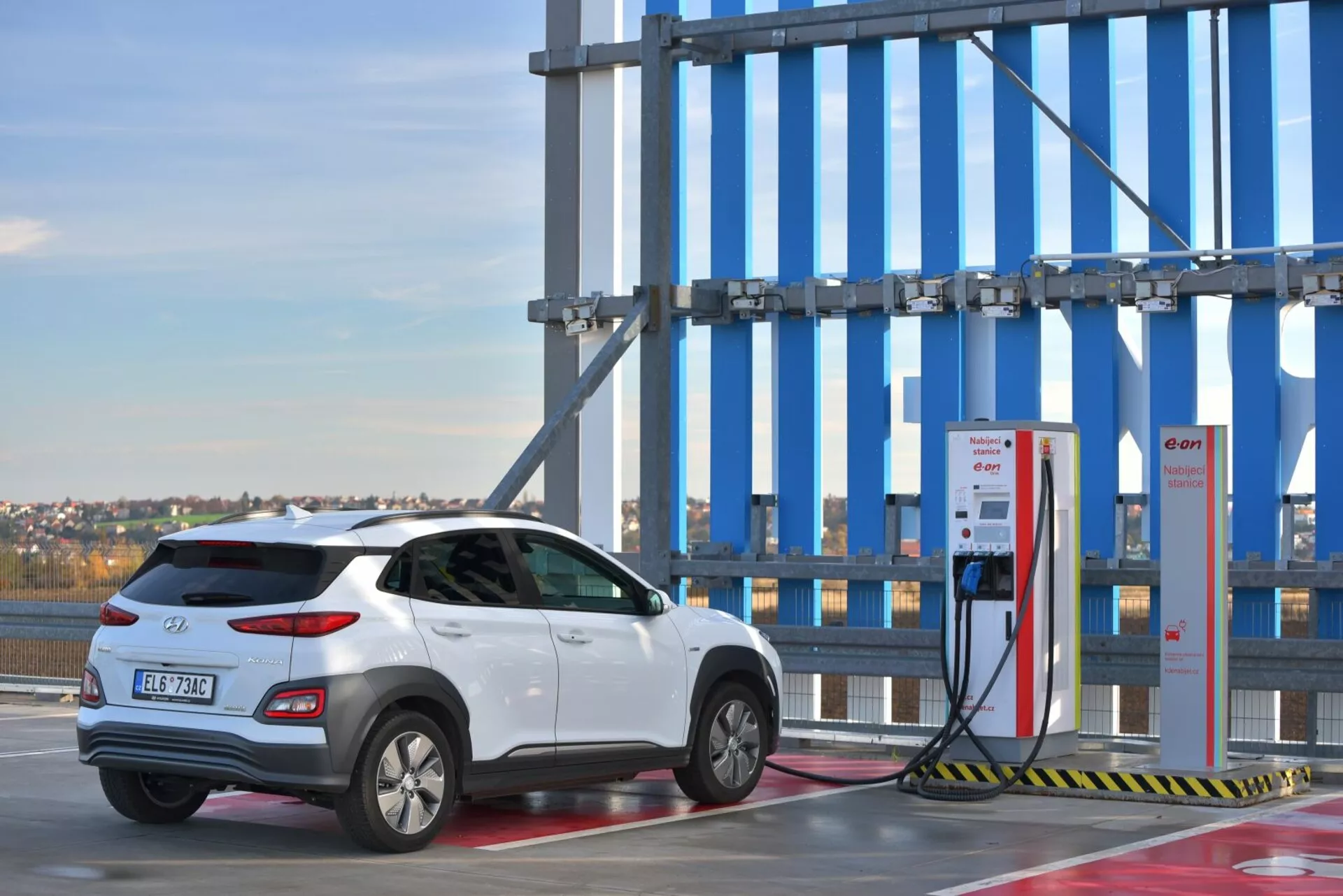 2021-es fehér Hyundai Kona elektromos SUV, háromnegyedes hátsó nézetben egy kültéri töltőállomáson, tiszta égbolt háttérrel