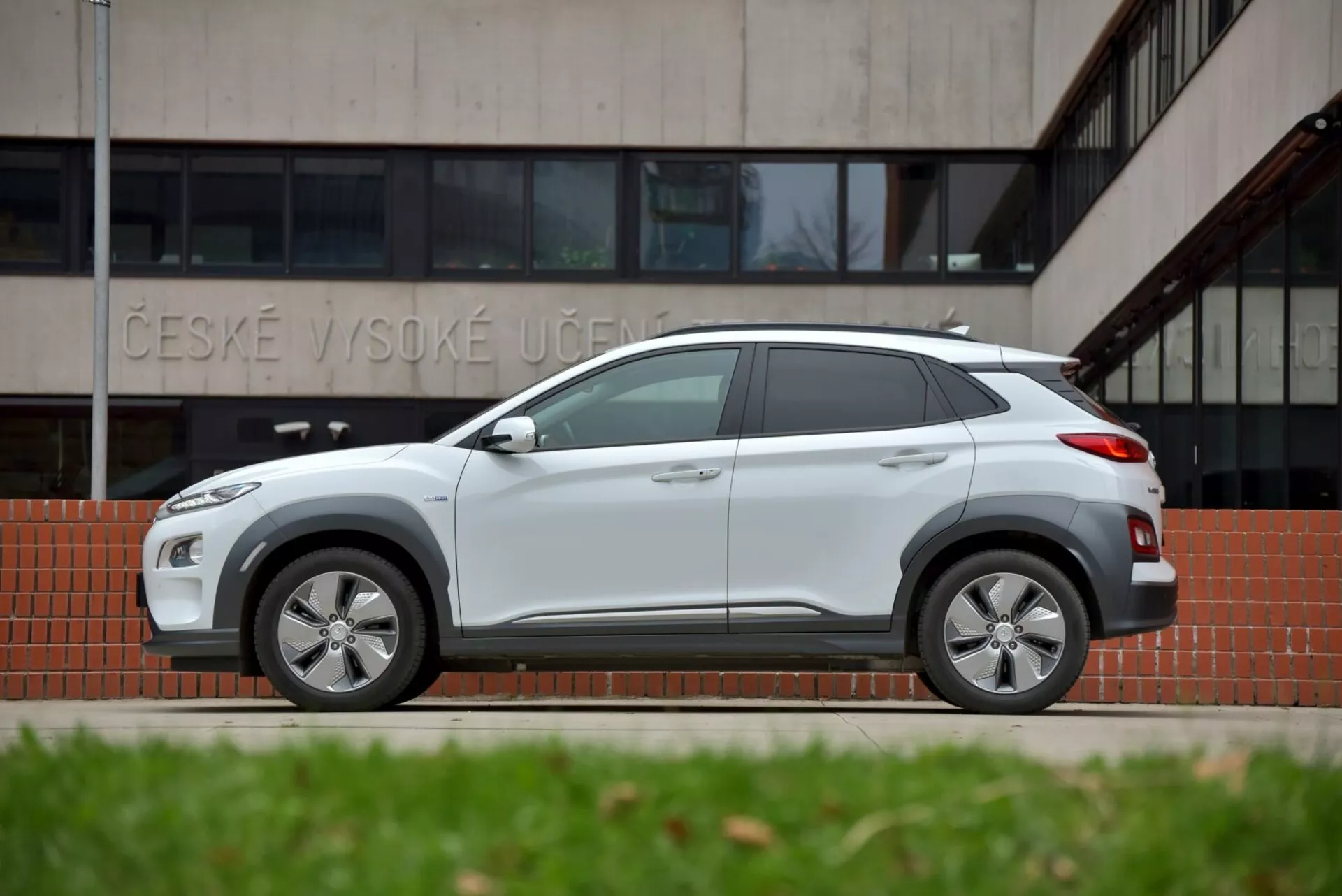 2021-es Hyundai Kona Elektro oldalnézete, kiemelve a világosszürke színét, elegáns kialakítását a karosszéria színű kerékívborításokkal és küszöbökkel, valamint modern jellemzőit, mint az ötvözet kerekek és cápauszony antenna, egy 'ČESKÉ VYSOKÉ UČENÍ' feliratú épület előtt, hangsúlyozva a városi és terepen való vezetésre való alkalmasságát