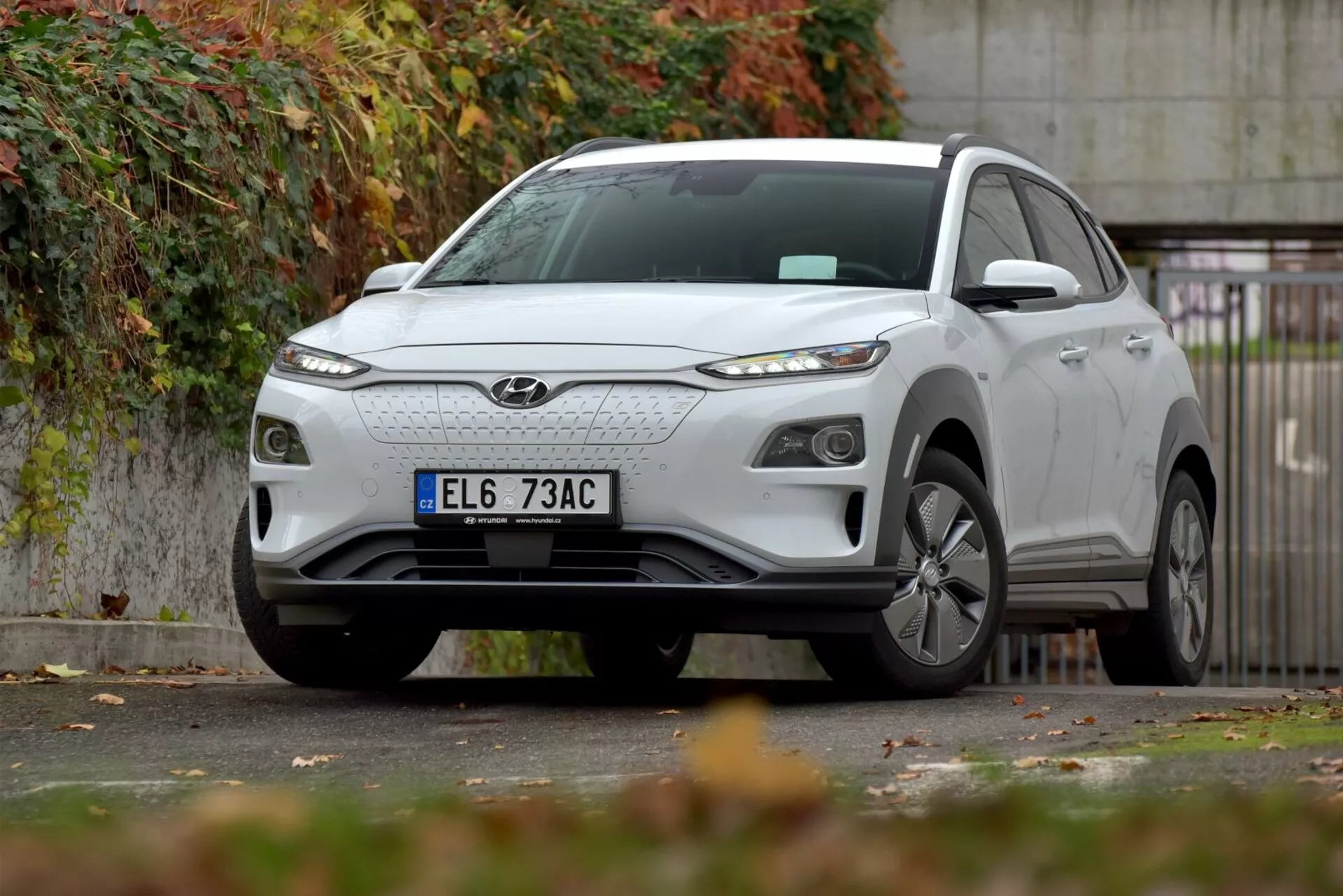 2021-es Hyundai Kona elektromos kompakt SUV parkol városi út szélén, bemutatva az első rácsot, elegáns LED fényszórókat, ötvözött kerekeket, és formált karosszériát az első bal oldal hangsúlyozásával, őszi táj közepette