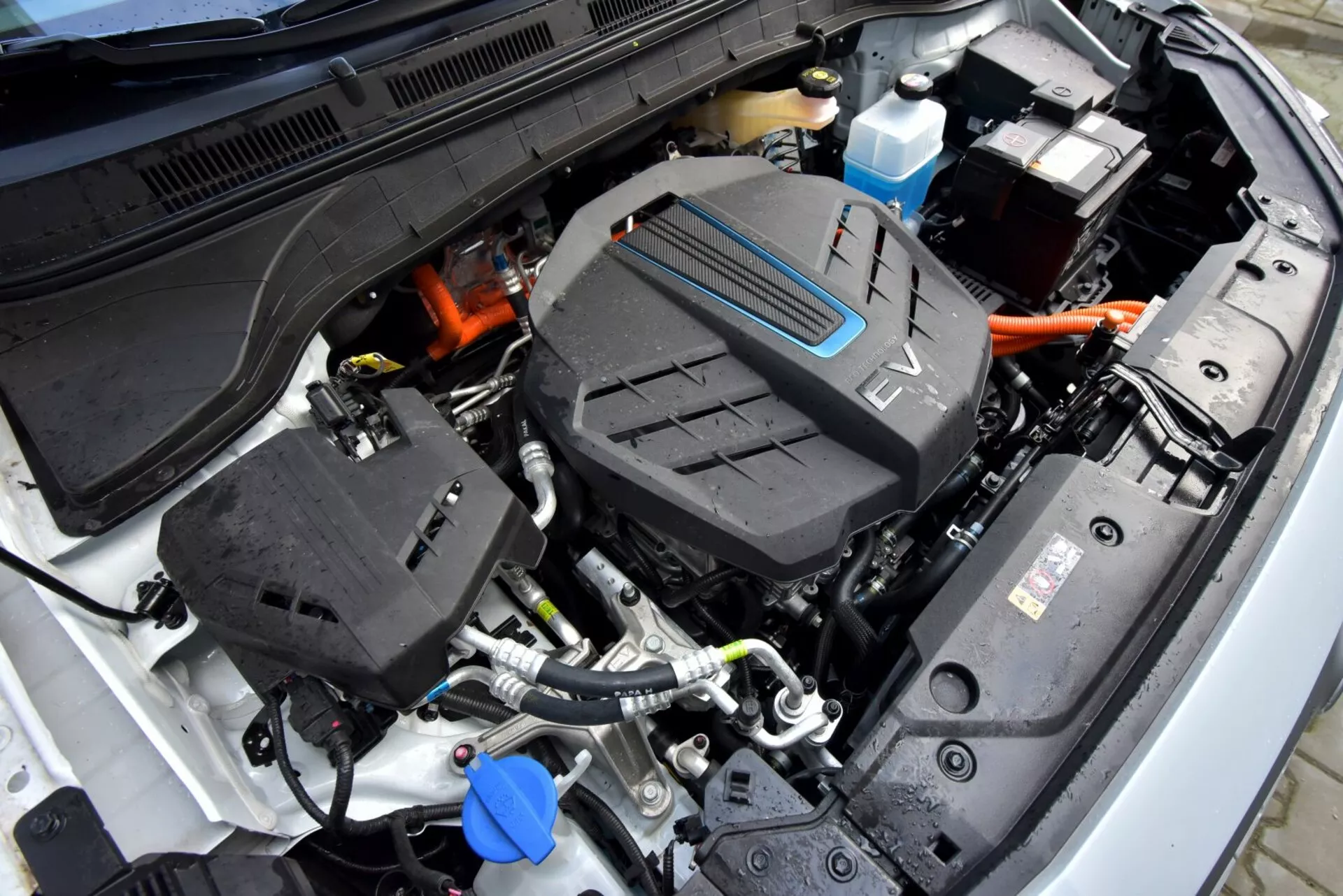 Részletes nézet a 2021-es Hyundai Kona Electric motorháztetőjéről, amely turbofeltöltős motor jelképet, légbeömlő rendszert, különböző folyadéktartályokat, nagyfeszültségű elektromos csatlakozókat és műanyag házas akkumulátort mutat be, előtérbe helyezve a fejlett autómérnöki technológiát.