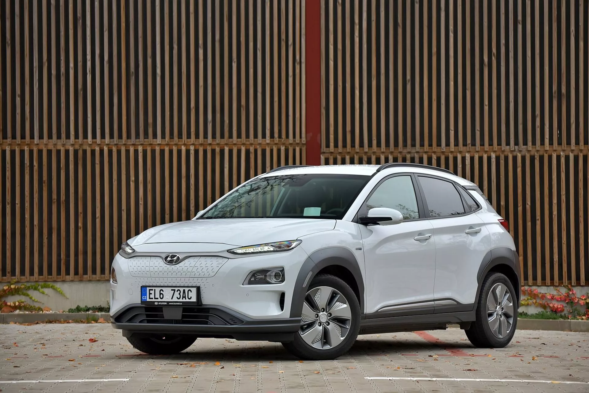 2021-es Hyundai Kona elektromos crossover SUV oldalnézeti képe, amely bemutatja az ötvözött kerekeket, a színre hangolt oldalsó tükrökön integrált irányjelzőket, négyajtós kialakítást, modern aerodinamikai vonalakat és íveket, LED fényszórókat, hátsó ablakokon sötétített üveget, minden évszakra alkalmas abroncsokat, és kortárs előlapi hűtőrácsot, járdán parkolva, hátterében egy fa homlokzatú épülettel.
