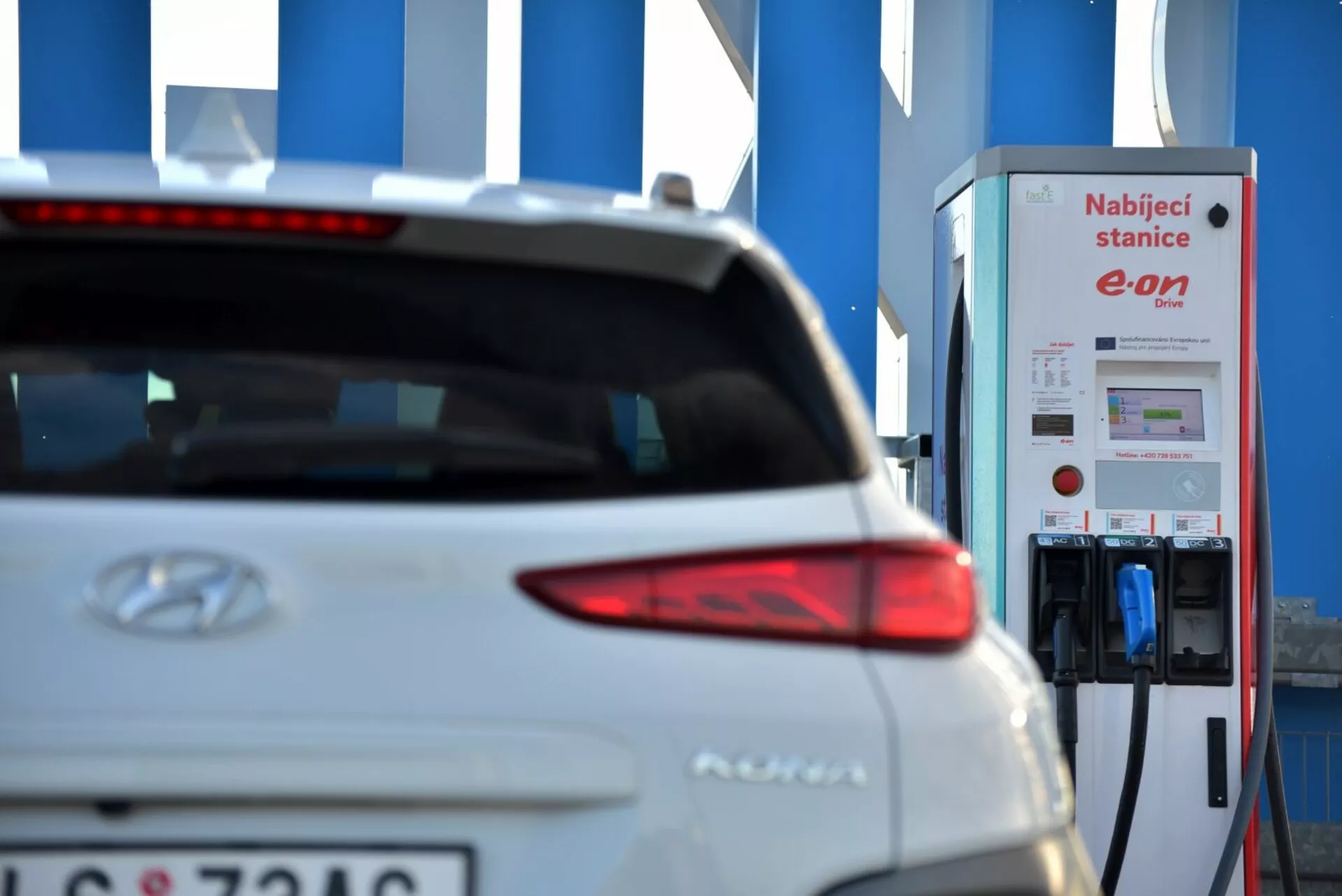 Közeli kép egy 2021-es fehér Hyundai Kona elektromos jármű hátuljának jobb oldaláról, a stílusos hátsó lámpa tervezésére összpontosítva, a Hyundai logó részben látható, és a modellnév a csomagtartó ajtó közepéhez közel. A csomagtartó ajtójának bal alsó részén részben látható rendszámtábla a magánszféra iránti odafigyelésre utal. A háttérben egy E.ON elektromos jármű töltőállomás található, kiemelve az autó elektromos jellegét egy elmosódott környezetben, utalva az elektromos mobilitásra és a fenntartható közlekedési technológiákra.