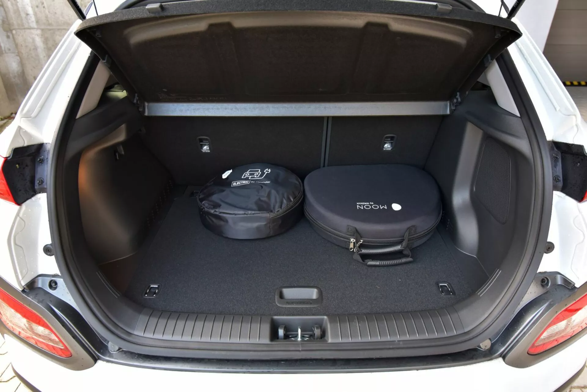 2021-es Hyundai Kona elektromos modell bemutatja a csomagtartó terét hátsó nézőpontból, a nyitott csomagtartóval, feltárva egy szőnyegezett rakodóteret csúszásgátló pályákkal és minimális tárgyakkal a tér méreteinek világos megtekintéséhez.