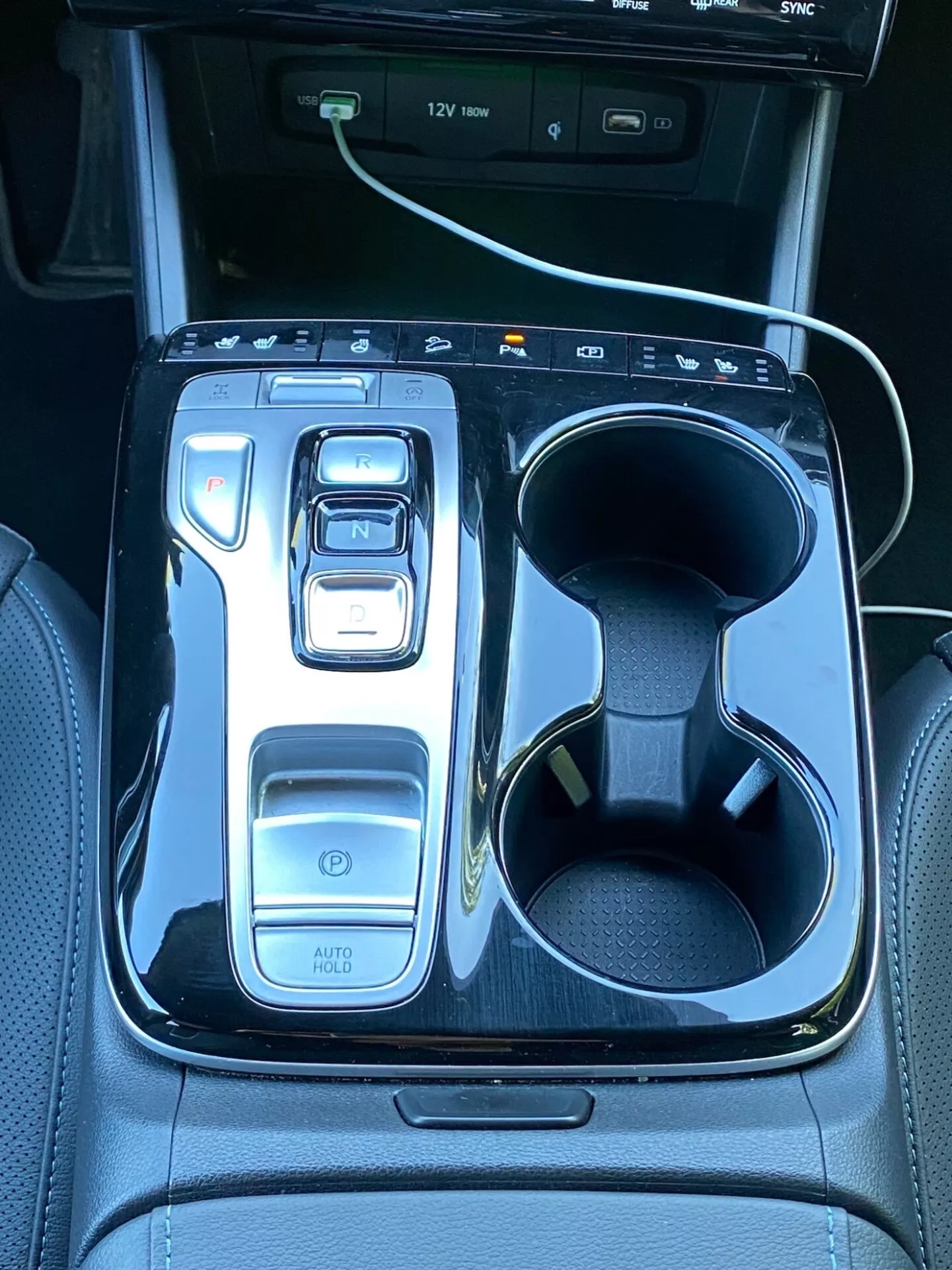 A kép egy 2020-as Hyundai Tucson középkonzolját ábrázolja, melyen egy elektronikus váltó kar látható 'N' állásban fényes fekete keretben. Balra találhatóak a vezérlők, beleértve egy bekapcsoló gombot, menetmód-választókat és az 'AUTO HOLD' funkciót. Jobbra kettős pohártartó, az egyikben fehér töltőkábel. A felső részen betekintést nyerhetünk egy infotainment kijelzőbe USB port jelzésekkel, ami a jármű technológiáját és luxusát jelzi.