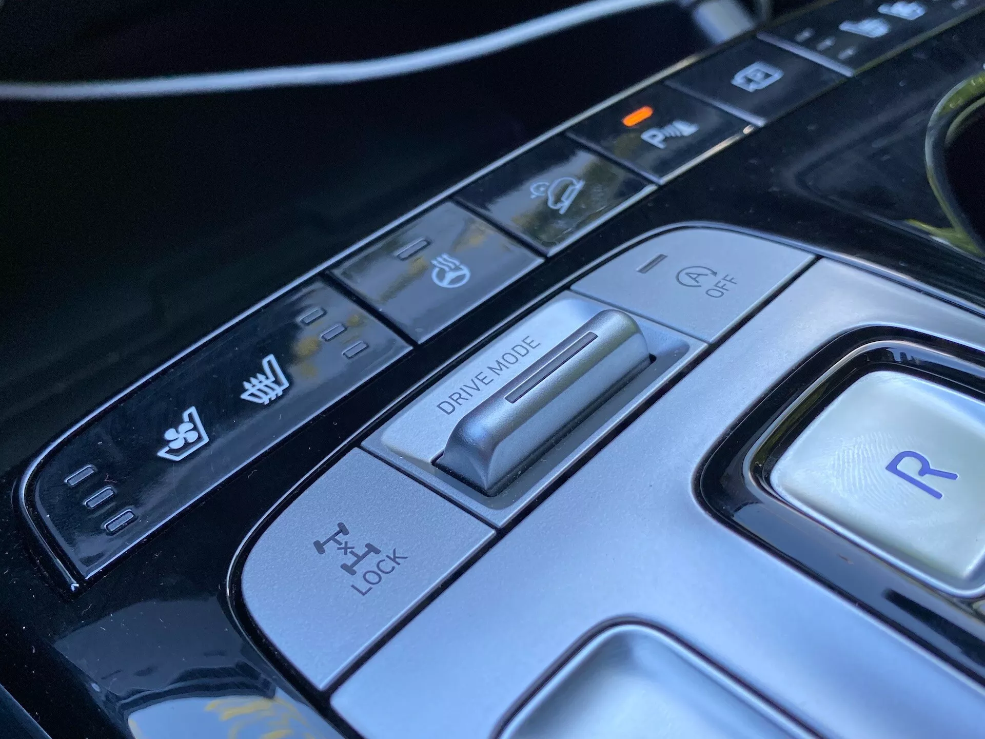 Közelkép a 2020-as Hyundai Tucson középkonzoljáról, amely a vezetési módválasztóra és a különböző vezérlőgombokra összpontosít. A kép egy fejlett vezetési módválasztót mutat különböző vezetési körülményekhez, egy kipörgésgátló panelt, egy visszameneti fokozatot jelző gombot és további vezérlőket a jármű stabilitásához és a parkolóasszisztens funkciókhoz. Az elegáns dizájn és a csúcstechnológiás anyagok egy kifinomult autóipari belsőt emelnek ki ennek a modern luxusautónak.