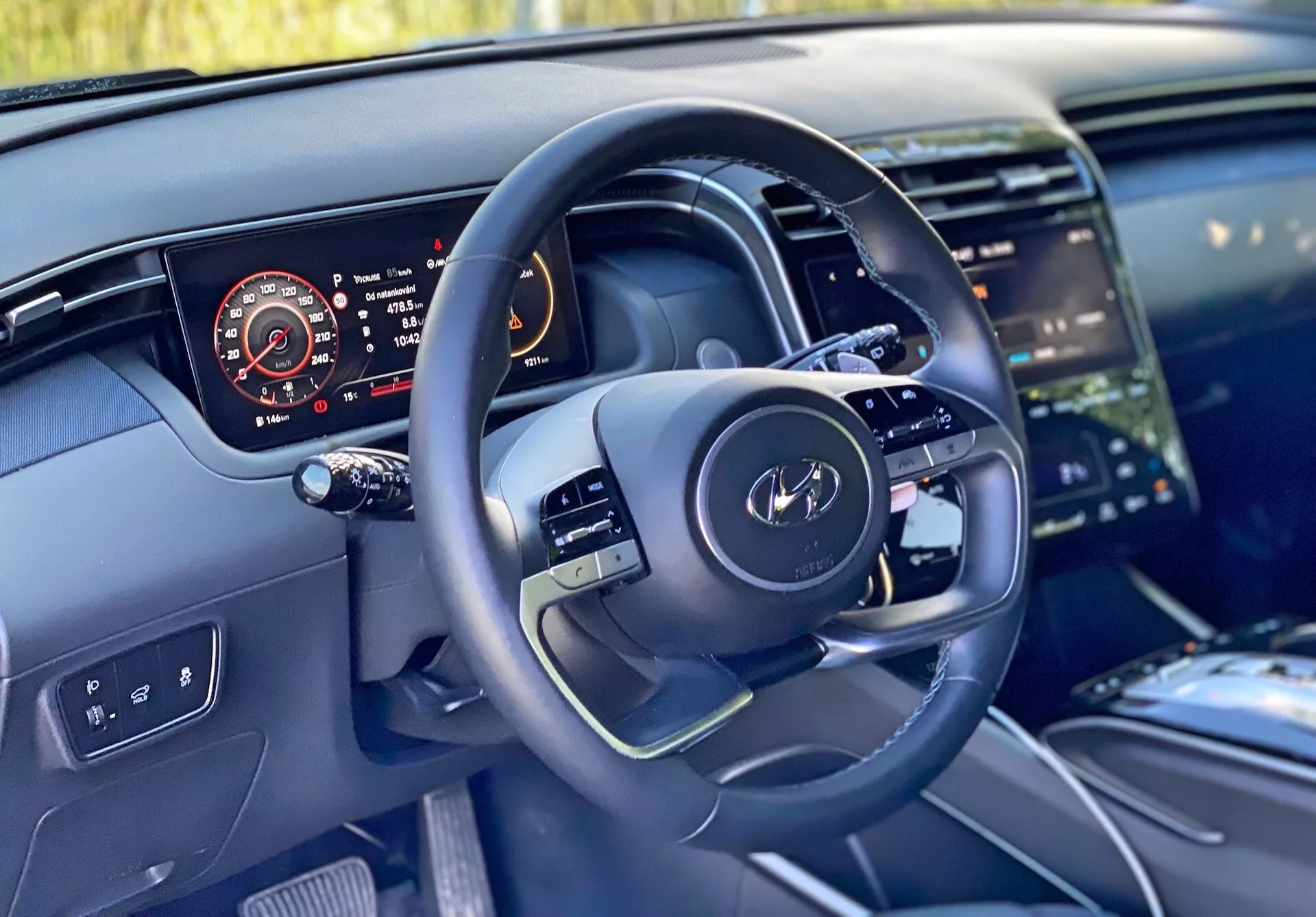 A Hyundai Tucson 2020 belső nézete a vezető szemszögéből, amely bemutatja a multifunkciós gombokkal ellátott modern kormánykereket és a digitális műszerfalat, amely a sebességmérőt, a fordulatszámmérőt, az üzemanyagszintet és a motor hőmérsékletét mutatja. A kép kiemeli a középkonzol egy részét is nagy érintőképernyős infotainment rendszerrel, a tiszta műszerfal dizájnt környezeti világítással és magas minőségű anyagokat, amelyek luxus kidolgozást sugallnak.