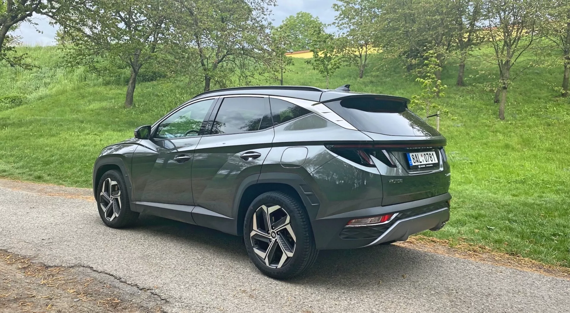 Háromnegyedes hátsó perspektíva egy 2020-as Hyundai Tucson 1.6 T-GDi Pure SUV-ról, amely egy keskeny úton áll egy vidéki környezetben, nagy, többrészes könnyűfém kerekeket, sötét fémes festést lejtős tetővonallal, hátsó légterelővel, hátsó ablakokon sötét üvegekkel, modern hátsó lámpákkal és kettős kipufogóvéggel, amely a hátsó lökhárítóba van beépítve.