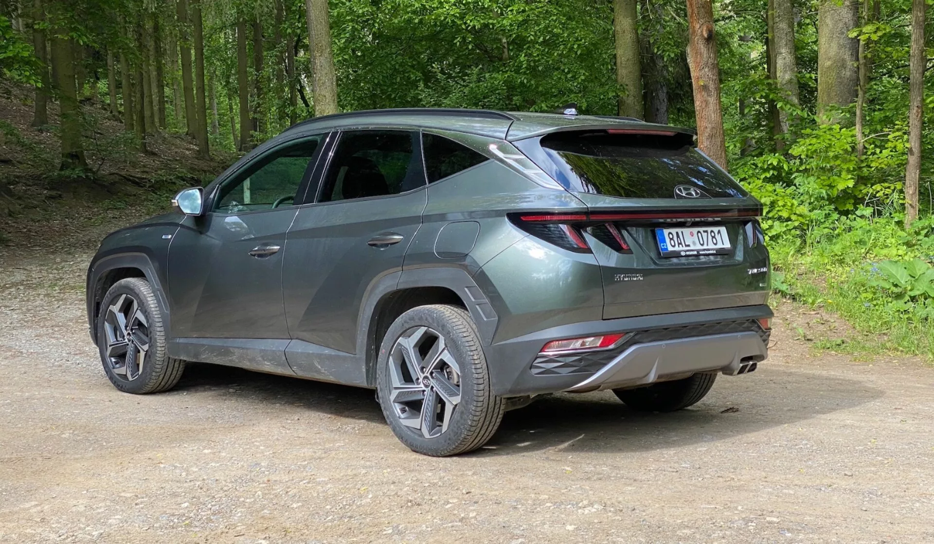 Egy modern SUV egy erdei területen lévő földúton parkol, egy enyhén megemelt hátsó háromnegyedes nézetből. A jármű elegáns, korszerű dizájnnal rendelkezik éles vonalakkal, hosszúkás hátsó lámpákkal, amelyek hátulról az oldalra húzódnak, látható spoilerrel, és a hátsó lökhárítón visszaverőkkel, ami fejlett biztonsági funkciókat sejtet. A fémes szürke jármű többküllős felnikkel és közúti abroncsokkal rendelkezik, fényes felülete alapján új vagy jól karbantartott. Az autó körül zöld, buja erdő van, a talaj tömörített földből és laza kavicsból áll.