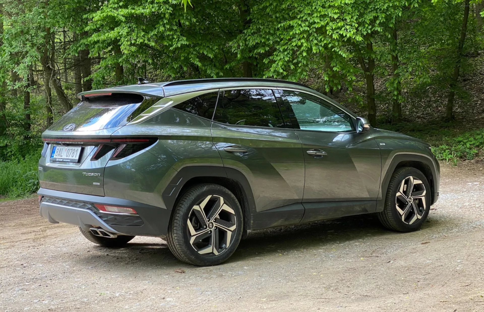 Háromnegyedes hátsó nézet egy modern 2020-as Hyundai Tucson SUV-ről, amelynek oldalsó profiljára és hátsó kialakítására összpontosít. A jármű balra van elhelyezve, a fotót jobbról készítették, kiemelve a jobb hátsó sarkot. A jellegzetes LED-es hátsó lámpák hátulról az oldalakra húzódnak, és egy tetőre szerelt spoiler sportos megjelenést kölcsönöz. A felfelé ívelő övvonal, a hátrafelé lejtő tetővonal, a sötétített ablakok és a karosszériával megegyező színű ajtófogantyúk kupészerű megjelenést kölcsönöznek neki. A nagy többrudas kialakítású ötvözet felnik és az alacsony profilú gumik megerősítik a luxus kinézetet. Az apró részletek, mint például a hátsó ablakmosó és a kerékív burkolatok, a praktikusságot és a robusztusságot jelzik. Az autó kavicson áll egy természetes környezetben, fákkal a háttérben.