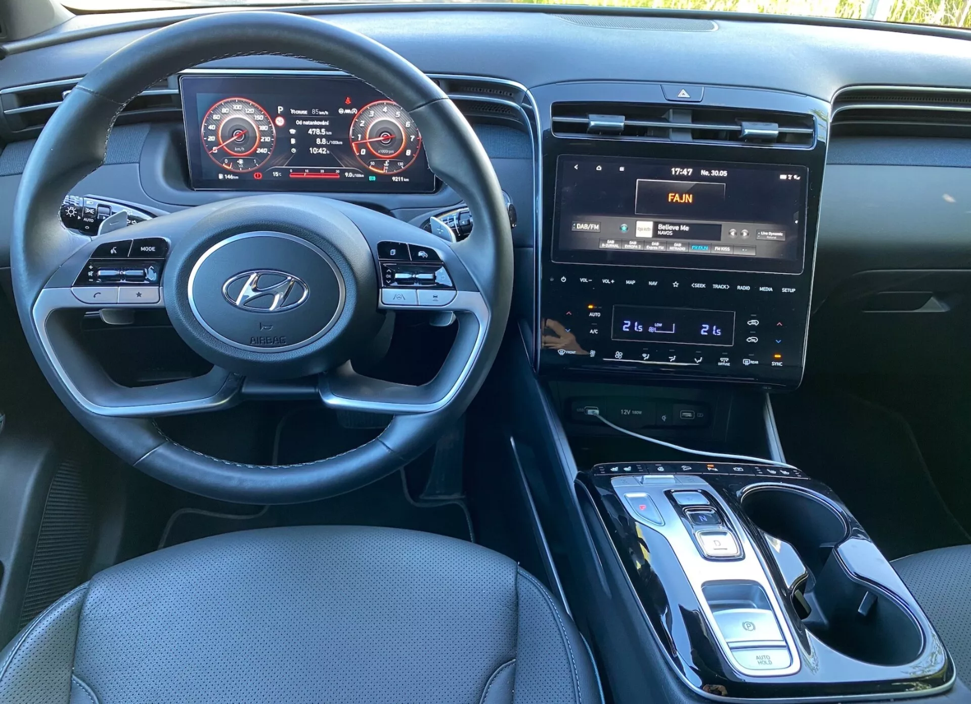 A 2020-as Hyundai Tucson belső nézete a vezetőülésből, bemutatva a Hyundai logóval ellátott kormánykereket és az audió, telefon és tempomat beállításokhoz használt kezelőszerveket. A műszerfal digitális kijelző panellel van felszerelve, piros kiemelt sebességmérővel és fordulatszámmérővel. A középkonzol infotainment-rendszert tartalmaz érintőképernyővel, amely légkondicionáló vezérlőket jelenít meg, fizikai gombokkal és tárcsákkal az éghajlati beállításokhoz, és egy automata váltóval elektronikus kiválasztóval. Látható egy potenciális vezeték nélküli töltőpad, sötét szövetkárpitozás és jól megvilágított kabin.