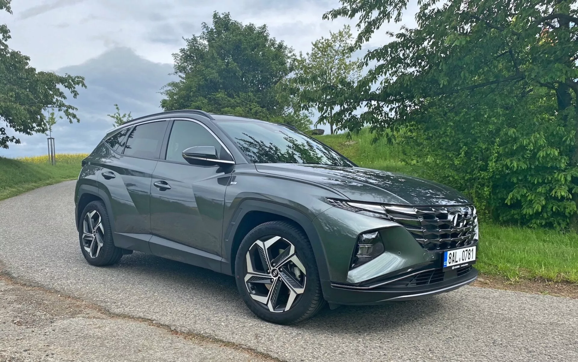 A képen egy modern Hyundai Tucson 1.6 T-GDi Pure SUV látható, amely egy aszfaltozott úton parkol, természetes háttérrel. Az autót egy elülső háromnegyedes szögből fotózták, így látható a feltűnő hűtőrács, az elegáns fényszórók, az első lökhárító ködlámpákkal vagy légbeömlőkkel, a nagy, többküllős alufelni és az oldalsó tükrök. Az SUV sziluettje hátrafelé lejt, ami sportos, aerodinamikus dizájnt sugall. Az enyhe nappali fényviszonyok között készült fotó kiemeli a jármű színét és részleteit erős árnyékok nélkül.