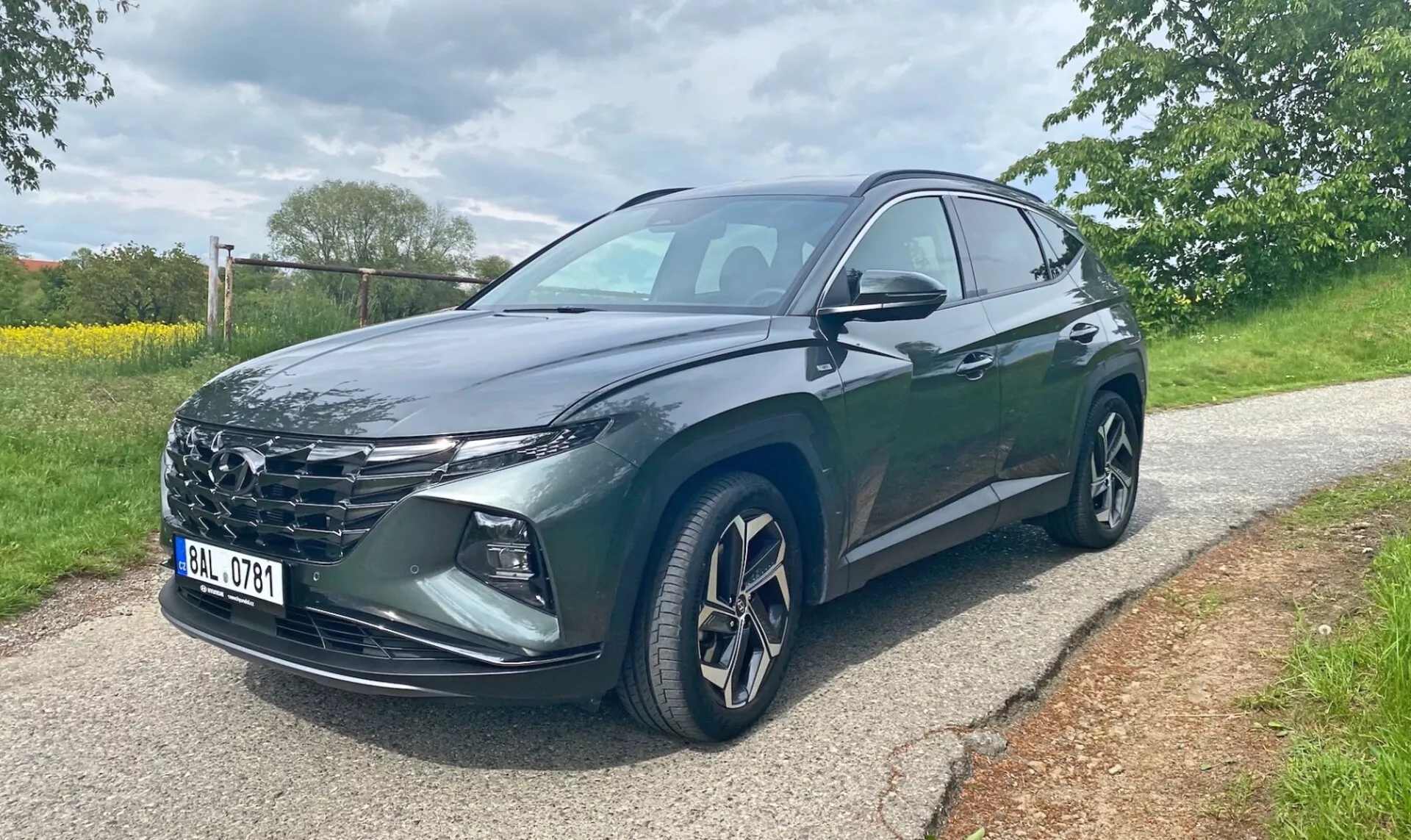 Hyundai Tucson 2020 1.6 T-GDI Pure SUV háromnegyed frontális szögből, burkolt úton állva, fák és fű látható a háttérben. A kép bemutatja a jármű hűtőrácsát, éles LED fényszóróit, ötvözött kerekeit, beépített irányjelzős oldalsó tükreit és sötét fémes fényezését. A vezető oldali első és hátsó ajtók láthatók. A fénykép napszaki felhős megvilágítási viszonyok között készült.