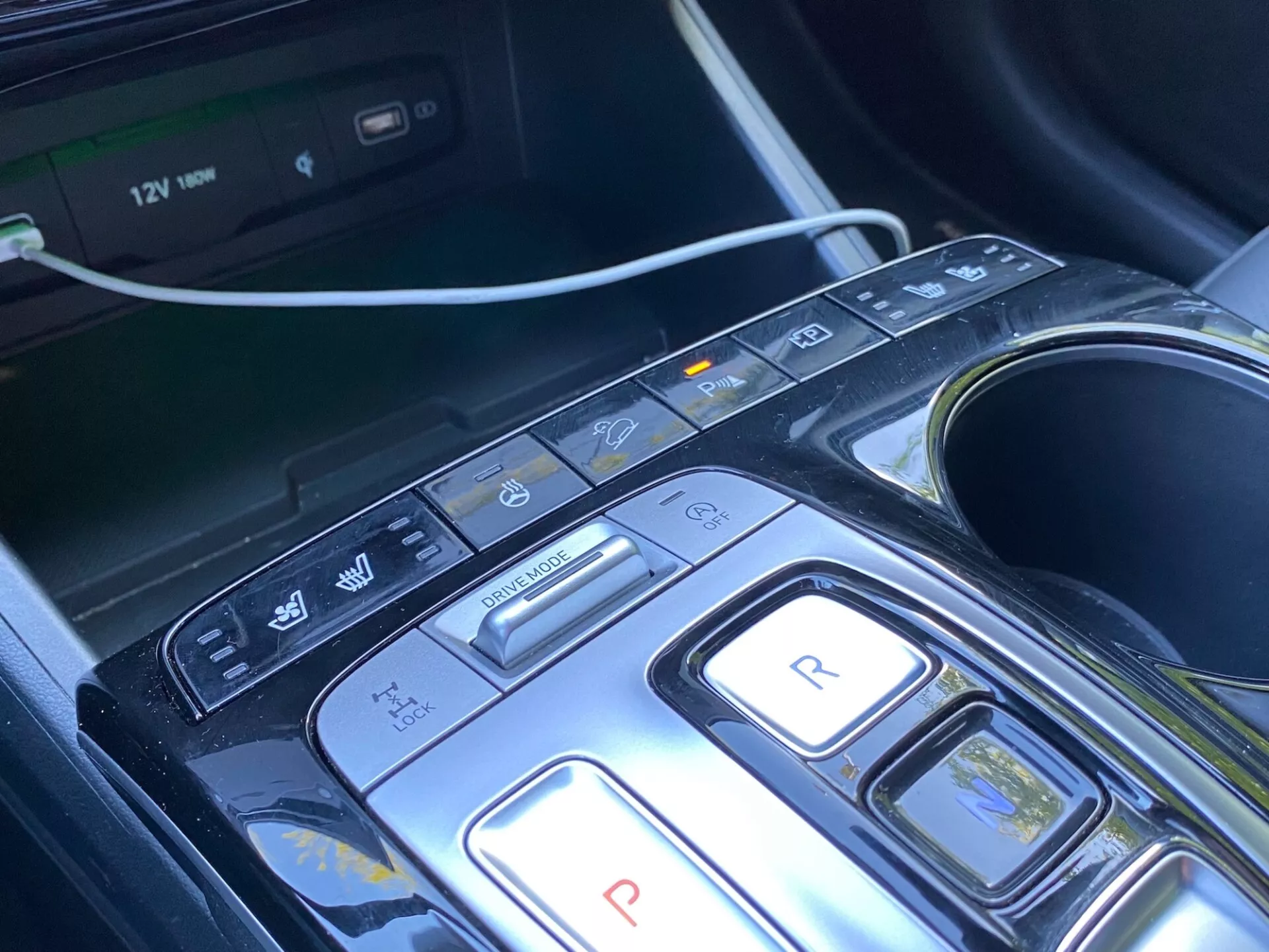 A 2020-as Hyundai Tucson 1.6 T-GDi Pure központi konzoljának és sebességváltó területének közeli képe, modern dizájn gombokkal a parkoláshoz, hátramenethez, neutrálishoz, vezetéshez és fék/regeneratív módokhoz, valamint további gombokkal az automatikus indítás/leállítás kikapcsolásához, parkolási érzékelőkhöz/automatikus parkoláshoz és sávmegtartás/sávelhagyási figyelmeztető funkcióhoz. A rendszer multimédiás képernyő, pohártartók és 12V-os aljzat jelenléte kényelemre, kényelmre és fejlett vezetői asszisztencia rendszerekre (ADAS) utal. A konzol elegáns dizájnnal, fényes fekete anyaggal és fémes elemekkel rendelkezik, ami luxus megjelenést sugall.