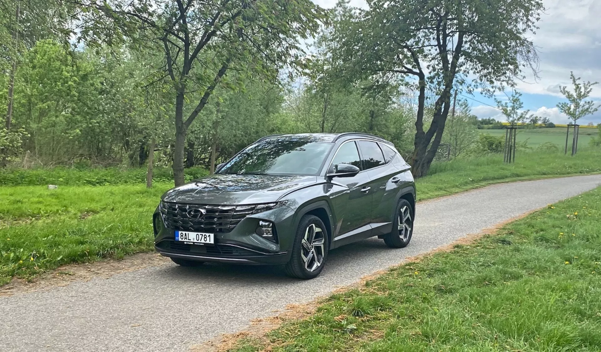 Háromnegyedes elülső nézet egy Hyundai Tucson 2020 crossover SUV-ról, amely egy burkolt úton parkol zöld háttérrel, bemutatva a jármű jellegzetes első rácsát, elegáns LED fényszóróit, nagy, többrudás könnyűfém kerekeit, hátrafelé lejtő tetővonalát, magas hasmagasságát, integrált oldalsó tükröket, sötétített üvegeit és stilizált karosszéria kontúrjait.