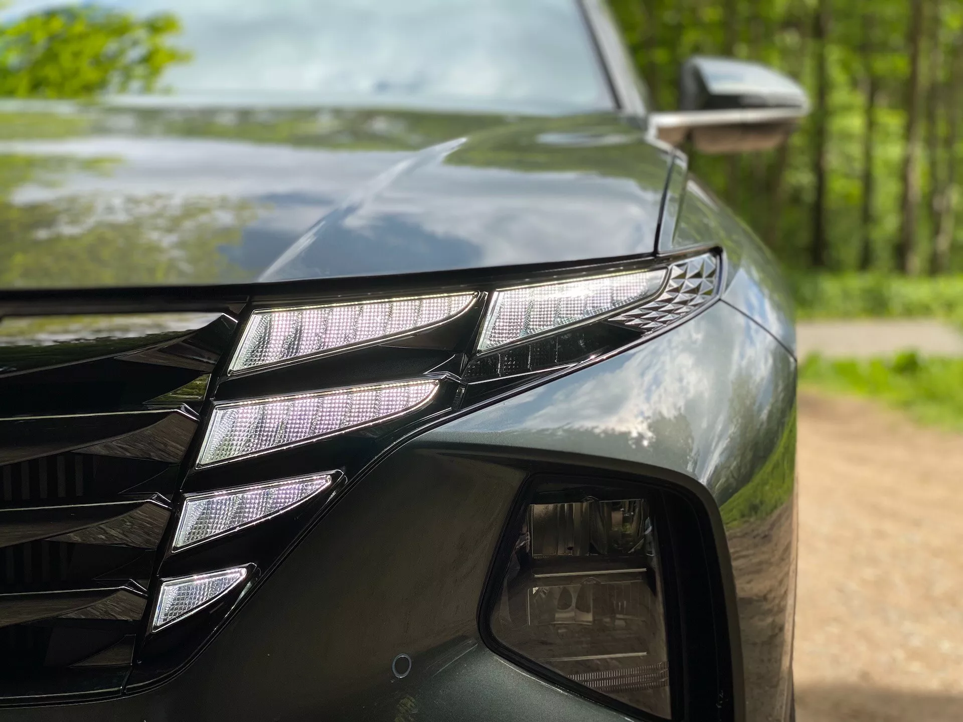 Közelkép a Hyundai Tucson 2020 (Hyundai-Tucson-1-6-T-GDi-Pure) bal első részéről, amely bemutatja az egyedi LED projektorokkal és jellegzetes LED DRL csíkkal ellátott LED fényszóró egységet. A sötét, fényes festék visszatükrözi a környező fákat. Látható a rács szögletes vonalaiból egy rész és az oldalsó tükör irányjelzővel.