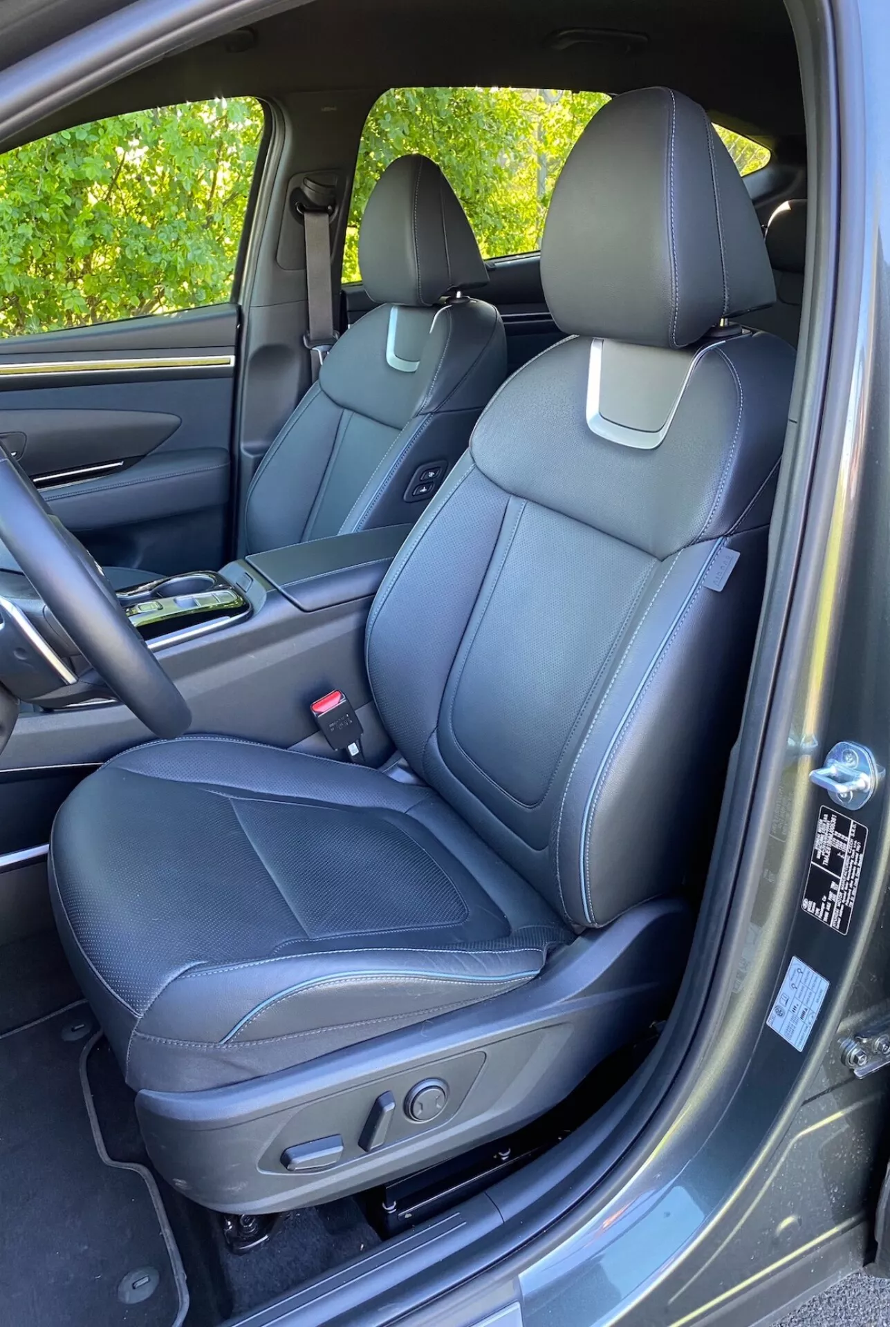 2020-as Hyundai Tucson 1.6 T-GDI Pure belső tere, mely bemutatja az első utasülést fekete bőrkárpitozással, részletes varrással és elektromos állítási vezérlőkkel. Látható középkonzol pohártartóval, ajtópanel passzoló anyagokkal, fogantyú, tárolórekesz és hangszórórács, amely fejlett audiorendszert sugall. Részben látható műszerfal és kesztyűtartó. Szőnyegszerű padlószőnyeg és enyhe napfényvisszaverődés az ajtópanelen.