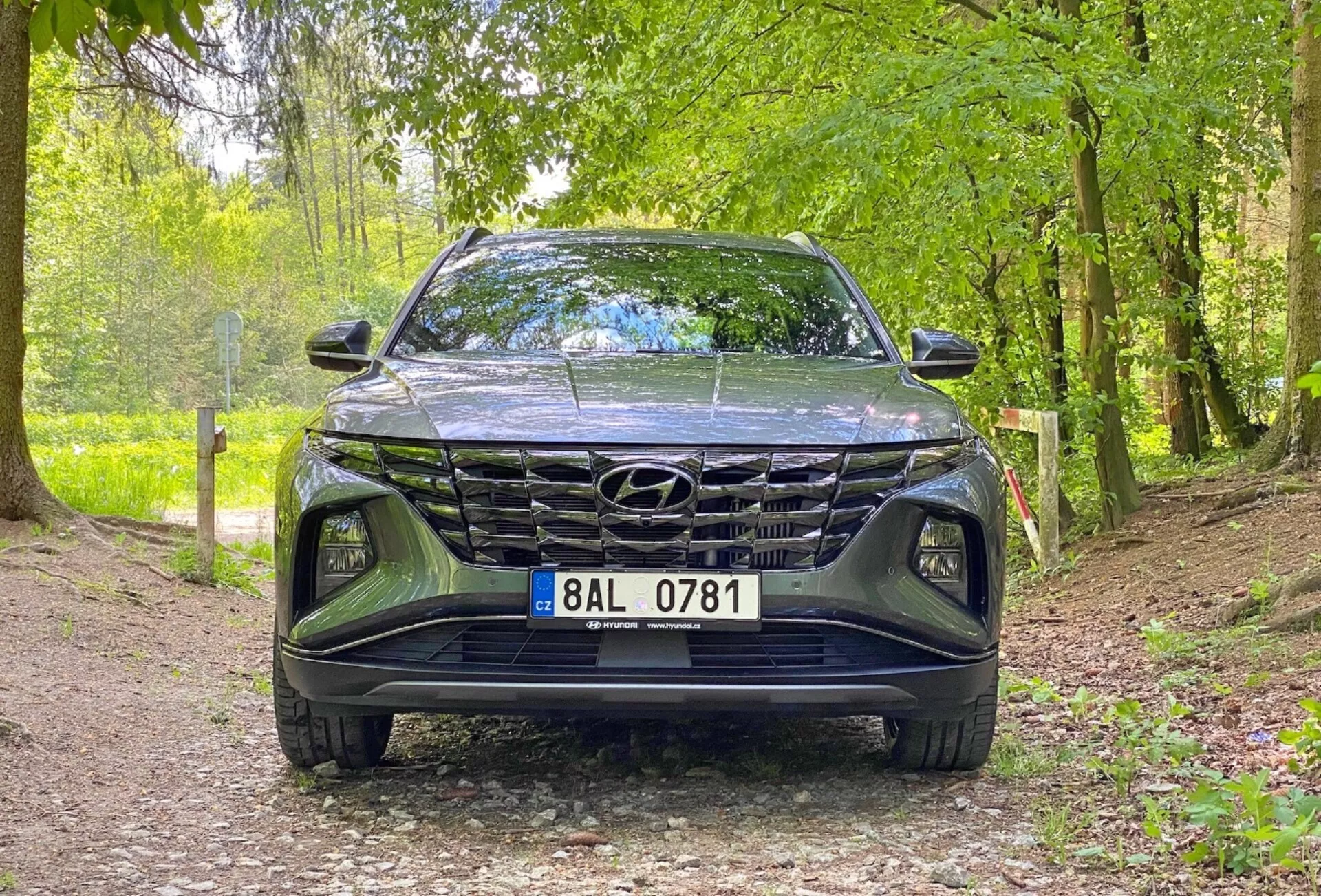 A 2020-as Hyundai Tucson 1.6 T-GDi Pure jármű elölnézeti képe, alacsony szögből készült felvétel, amely a hűtőrácsra, az első lökhárítóra és a motorháztető területére összpontosít. Az autó egy kavicsos úton parkol természetes környezetben, ami azt sugallja, hogy alkalmas szabadtéri környezetekre. A fő jellemzők közé tartozik a markáns hűtőrács kialakítás, a karcsú LED fényszórók és a jól látható Hyundai embléma.