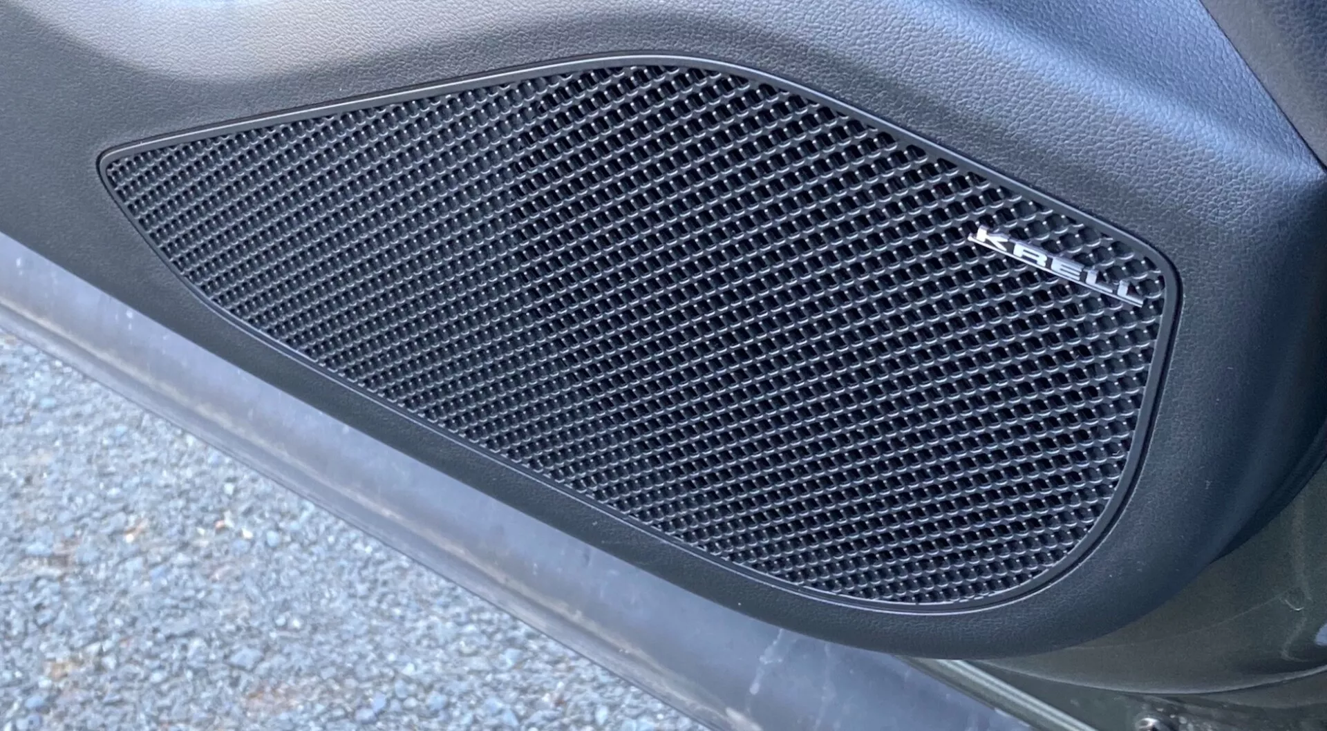 A Hyundai Tucson 2020 1.6 T-GDI Pure közelről mutatott oldalnézete, amely egy oldalrácsra összpontosít, amely rombusz alakú kivágásokból áll. A rács, amely egy öntött kontúrba van beillesztve, a jármű karosszériapaneljébe van építve matt felülettel, amelyet sima, ívelt felület vesz körül. A kép ferde szögből, alulról felfelé készül, természetes külvilágítással és aszfaltdarabokkal, út törmelékkel, ami fokozza az elegáns, teljesítményorientált megjelenést.