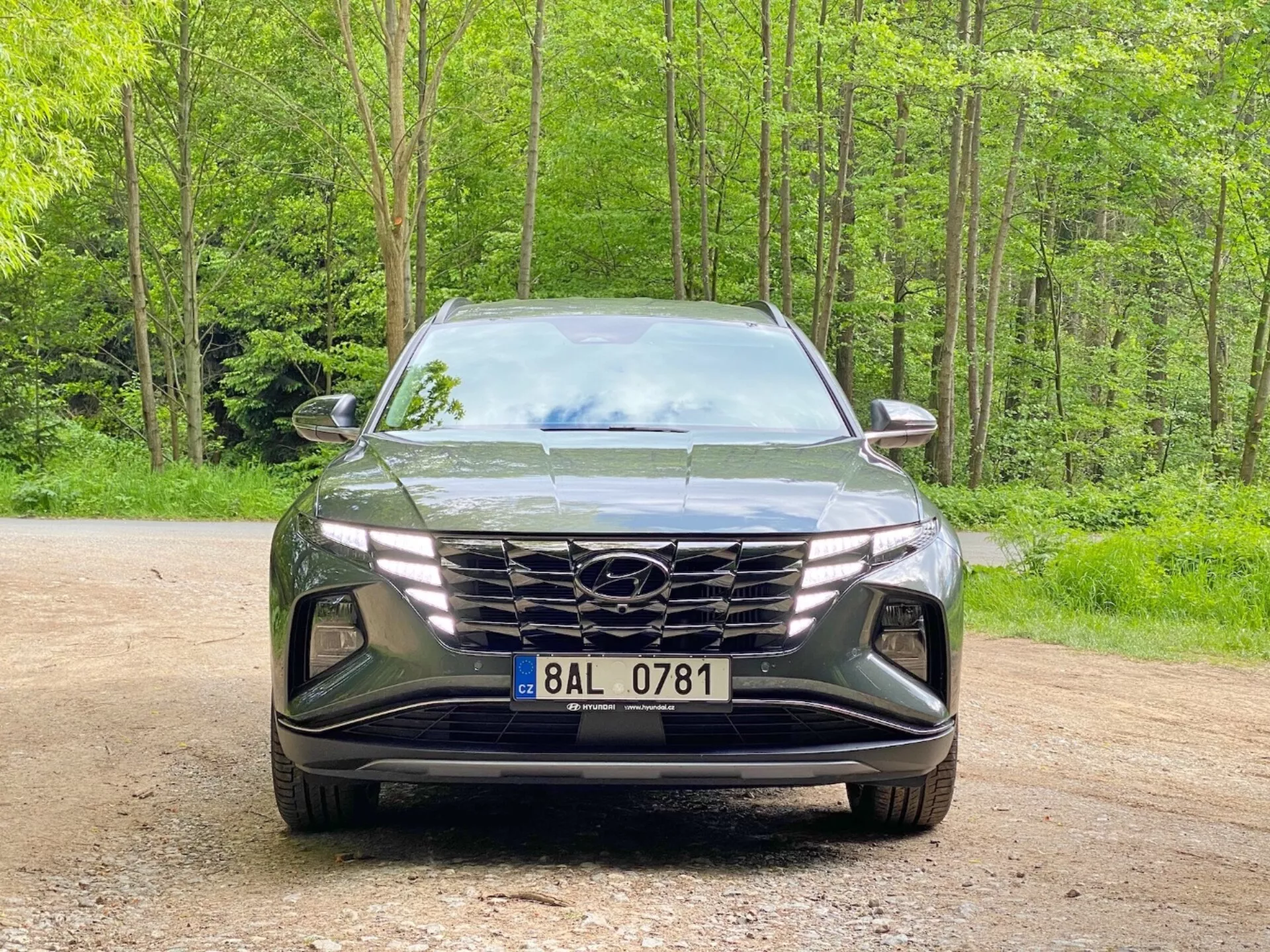 Elölnézeti kép egy Hyundai Tucson 2020 (Hyundai Tucson 1.6 T-GDI Pure) modern SUV-ról, amely egy murvás úton parkol, erdős háttérrel. A kép az elegáns, agresszív formatervezést ábrázolja, nagy geometrikus hűtőráccsal, amely a gyártó logóját tartalmazza, keskeny, szögletes LED-fényszórókkal és légbeömlőkkel valamint ködlámpa-fészkekkel díszített formázott első lökhárítóval, hangsúlyozva a fejlett aerodinamikát és világítási technológiát.