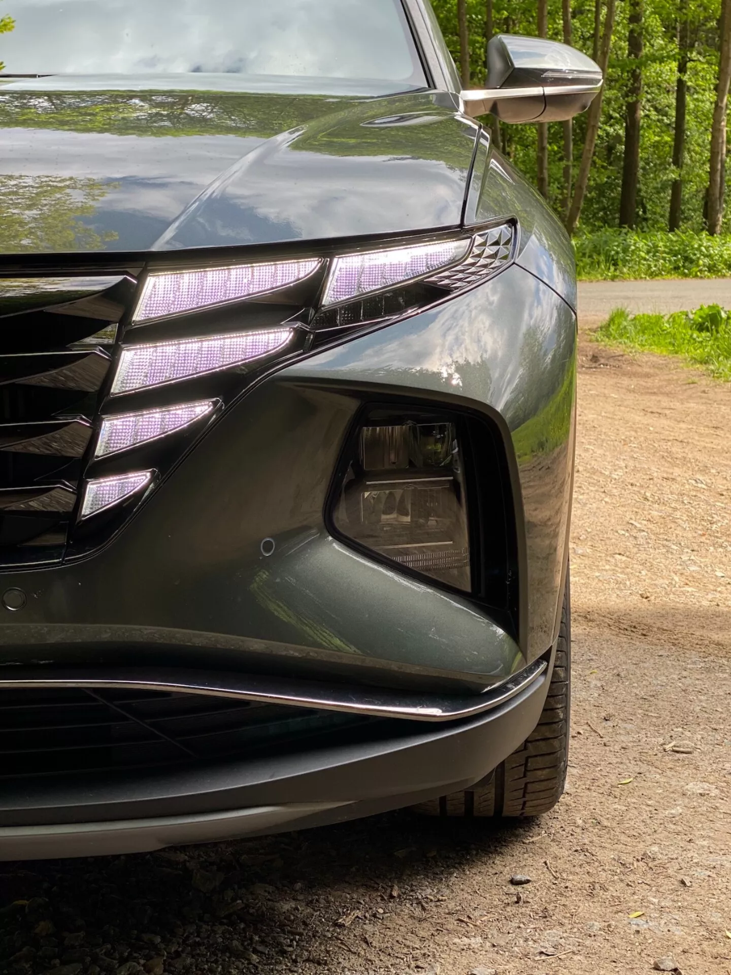 2020-as Hyundai Tucson 1.6 T-GDI Pure bal első sarkának közelről nézve, bemutatva a fényszórót LED nappali menetfénnyel és projektoros lencsével, az első rács egy részét vízszintes vonalakkal és króm díszítéssel, a levegőbeömlő részt ködlámpa házzal, sima vonalvezetéssel rendelkező sarkosított első lökhárítót és az integrált irányjelzőt a külső tükörben. A sötét, fényes festés kiemeli az autó aerodinamikai formáit.