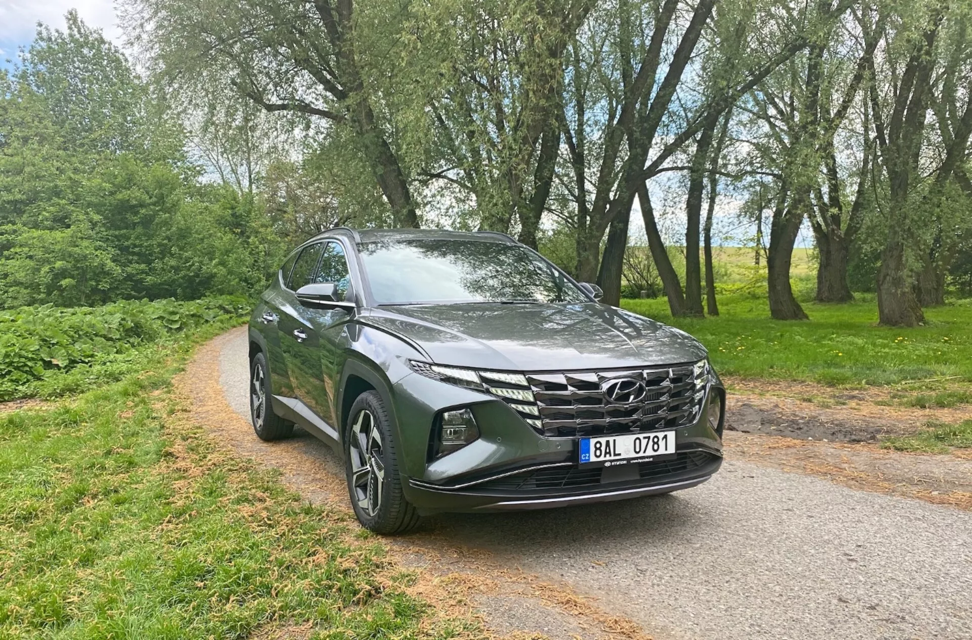 A 2020-as Hyundai Tucson 1.6 T-GDi Pure elölnézeti háromnegyedes képe, amely modern SUV jellemzőket mutat be, mint például elegáns LED fényszórók, jellegzetes geometriai elülső hűtőrács, aerodinamikus motorháztető, ötvözetből készült felnik, irányjelzőkkel integrált visszapillantó tükrök és fémes fényű festék, egy fás háttérrel szűk aszfaltozott úton, kiemelve sokoldalúságát és vonzerejét mind városi, mind vidéki környezetben.
