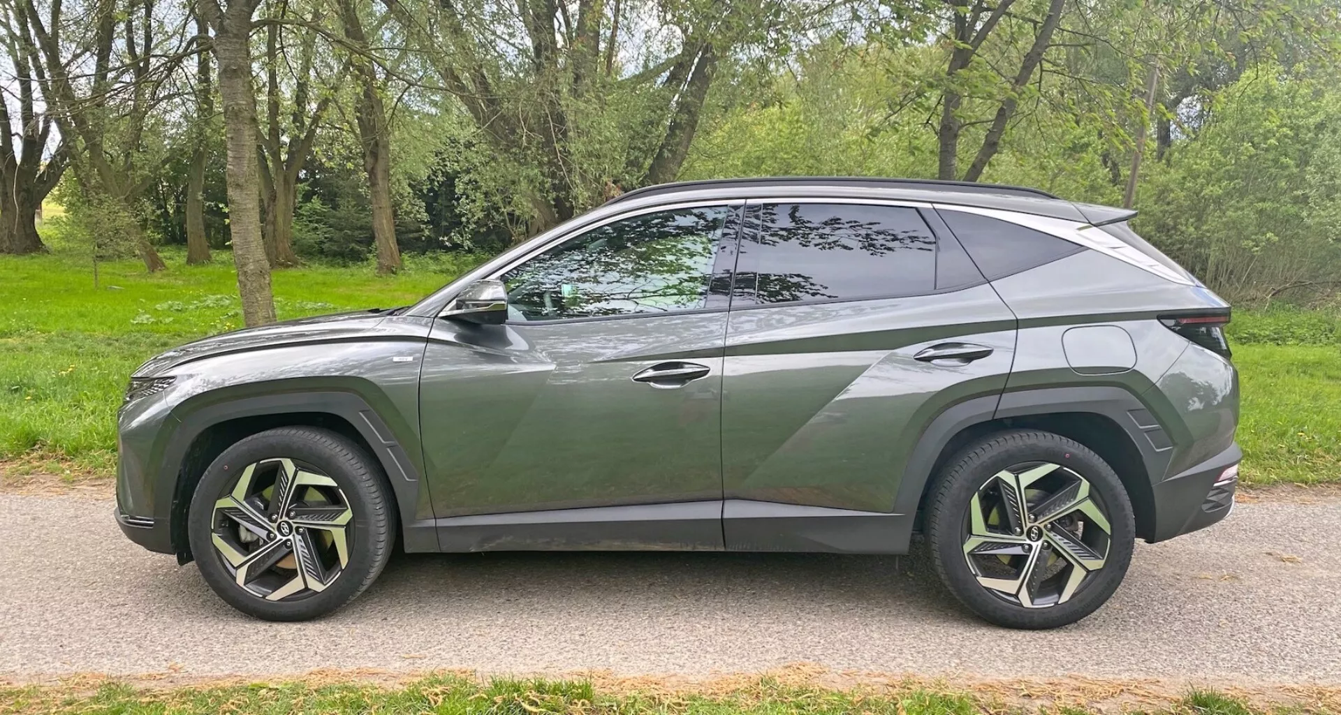 Oldalnézet egy modern Hyundai Tucson 1.6 T-GDI Pure 2020 SUV-ról, amely aszfaltozott úton, füves terület mellett parkol. Bemutatja az elegáns, aerodinamikus dizájnt lejtős tetővonallal, jellegzetes karosszéria vonallal, nagy, több küllős könnyűfém kerekekkel és alacsony profilú gumiabroncsokkal, sötétített ablakokkal, látható B-oszloppal, süllyesztett ajtófogantyúkkal, a karosszéria színéhez illeszkedő oldalsó tükrökkel és kromozott díszléccel az ablak szélén. A jármű modern stílust és potenciálisan fejlett funkciókat mutat oldalnézetből.