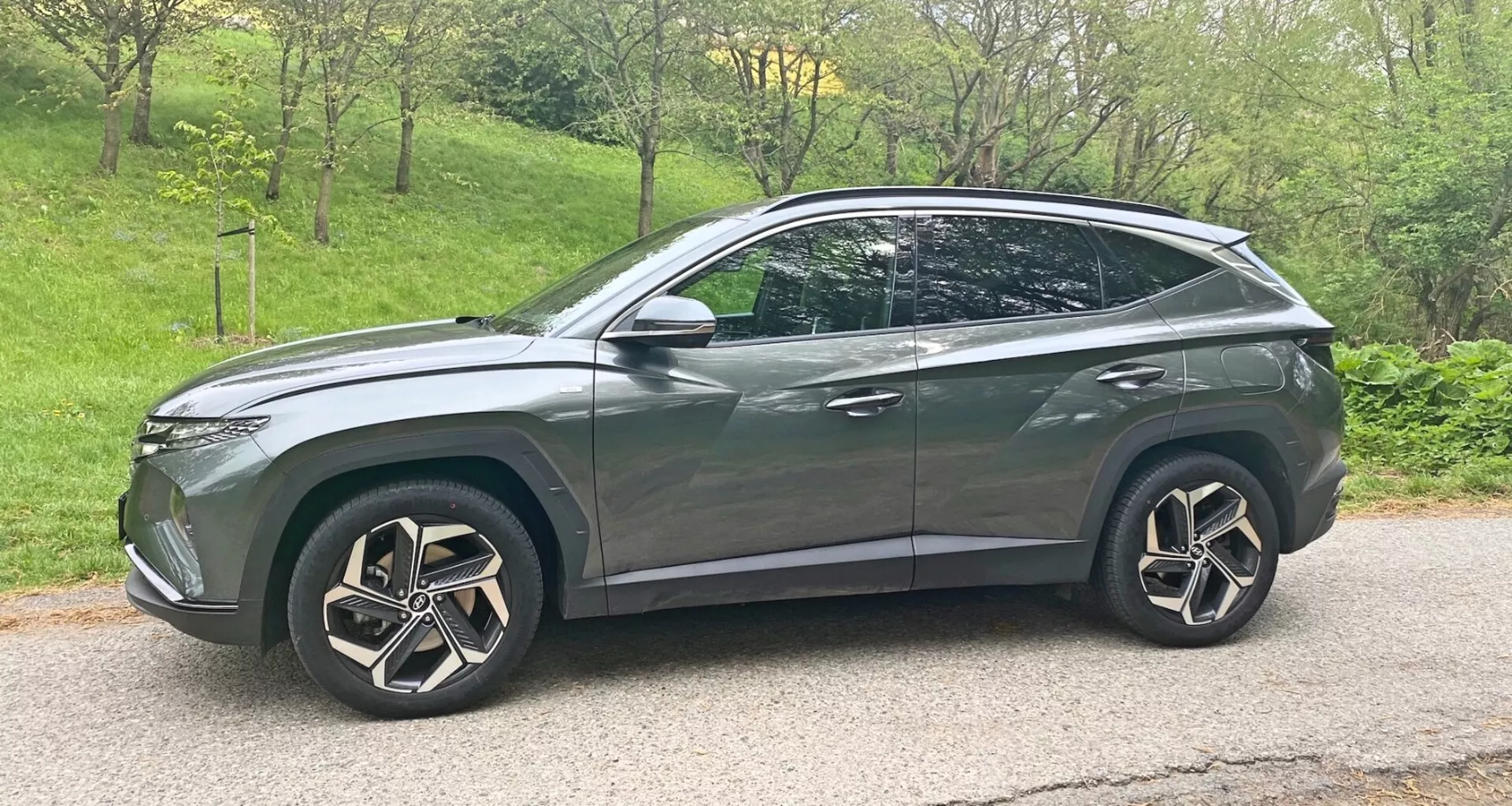 Egy modern 2020-as Hyundai Tucson 1.6 T-GDI Pure crossover SUV oldalsó profilképe fémes szürkében, fényes befejezéssel. A jármű nagy, bonyolult többküllős alufelnikkel, LED fényszórókkal, enyhén sötétített ablakokkal, áramvonalas karosszéria vonalakkal, amelyek lejtős tetővonalban végződnek hátsó spoilerrésszel, tetősínekkel, integrált irányjelzőkkel ellátott oldalsó tükrökkel, valamint króm vagy fémes díszítésekkel rendelkezik. A jármű egy aszfaltúton helyezkedik el enyhe lefelé irányuló szöggel, és a kép keretében inkább jobbra van elhelyezve.
