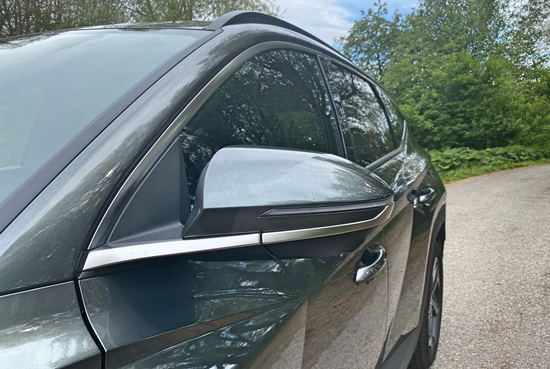 A 2020-as Hyundai Tucson 1.6 T-GDI Pure közelről megörökített oldalnézete, amely a modern dizájnt és technológiai jellemzőket emeli ki, a vezető oldalára fókuszálva. Az alacsony szögből készült fotó kiemeli az elegáns, aerodinamikai kialakítású oldalsó tükröt beépített irányjelzőkkel és fekete díszítéssel, fejlett funkciókat sugallva. Az elülső ajtóvezető oldali húzott kilincs kulcs nélküli beléptető rendszert jelez, hozzájárulva a jármű aerodinamikai hatékonyságához. A fényes, karosszéria színű ajtók és a krómmal díszített ablakkeretek prémium csomagot jeleznek. A háttérben látható elemek, mint például a fák és az ég, szabadtéri környezetet sugallnak.