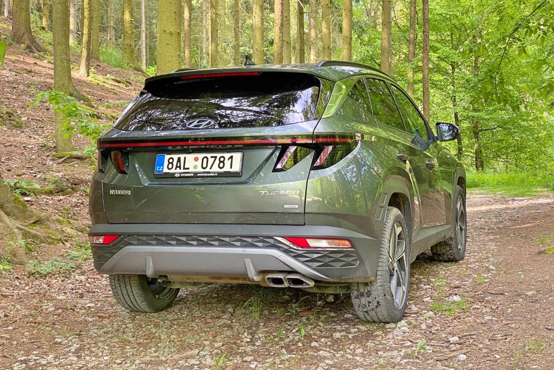 Kép egy modern Hyundai Tucson 2020 SUV-ról egy kavicsos erdei környezetben, hátulról háromnegyedes szögből felvéve. A fotó kiemeli a jármű jobb oldalát és hátsó profilját a látható hátsó ajtóval, LED-es hátsó lámpákkal, dupla kipufogórendszerrel, hátsó légterelővel és könnyűfém felnikkel.