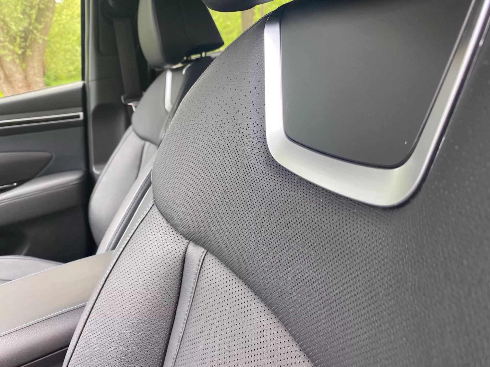 Közelkép a 2020-as Hyundai Tucson belteréről, a jobb oldali A-oszlopra és a jobb első ülés egy részére összpontosítva. Az A-oszlop texturált anyaggal van borítva, valószínűleg szövet vagy szintetikus bőr, ami esztétikai vagy zajcsökkentési célokat sugall. Az ülés kéttónusú kialakítású és szellőzés céljából perforált. Az A-oszlop és a tetőkárpit találkozásánál található jellemző egy meghatározó elem, amely a rendszer része lehet, légzsák komponens vagy vezetősegítő érzékelő, kiemelve az autó fejlett biztonsági vagy technológiai funkcióit.