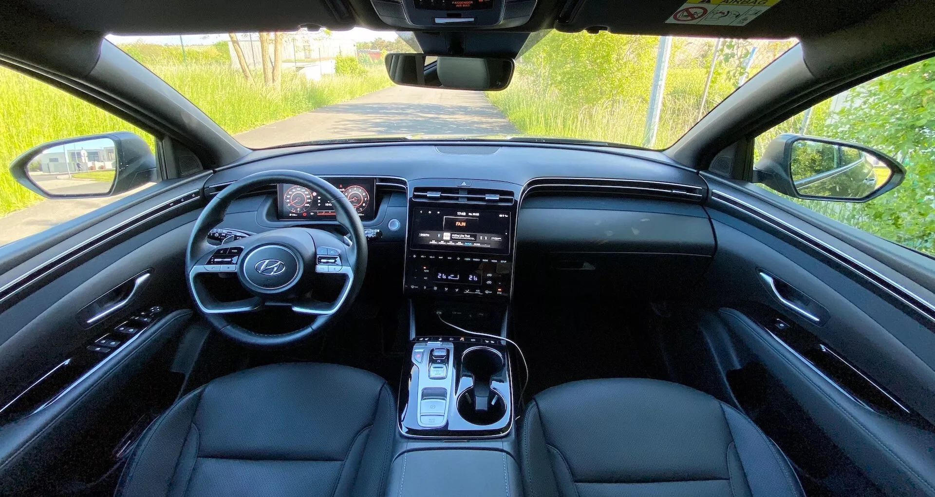 A Hyundai Tucson 2020 belső nézete a hátsó ülésből, bemutatva az elülső műszerfalat, a kormánykereket beépített vezérlőkkel, a digitális műszeregységet, a nagy érintőképernyős infotainment rendszert, a klímavezérlő gombokat, a sebességváltó kart és a bőrözött első üléseket természetes nappali fényben.