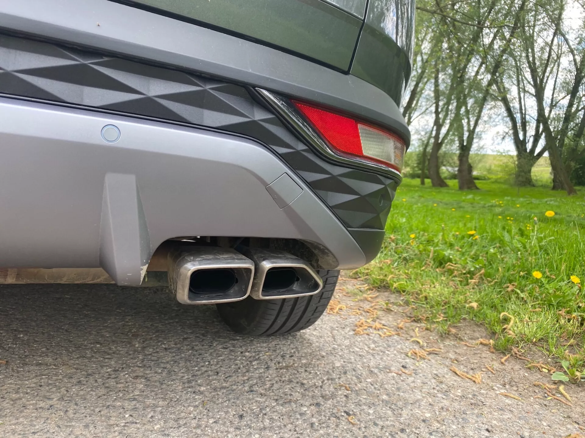 A 2020-as Hyundai Tucson hátulsó sarkának közeli képe, földszintes szögből. A látható részletek közé tartozik a kéttónusú hátsó lökhárító, egy részleges LED hátsólámpa és a fémes, kettős kipufogócsövek négyszögletes végekkel, ami sportos kivitelre utal. A jármű elegáns kialakítása és metálfényezése visszatükrözi a természetes környezetet.