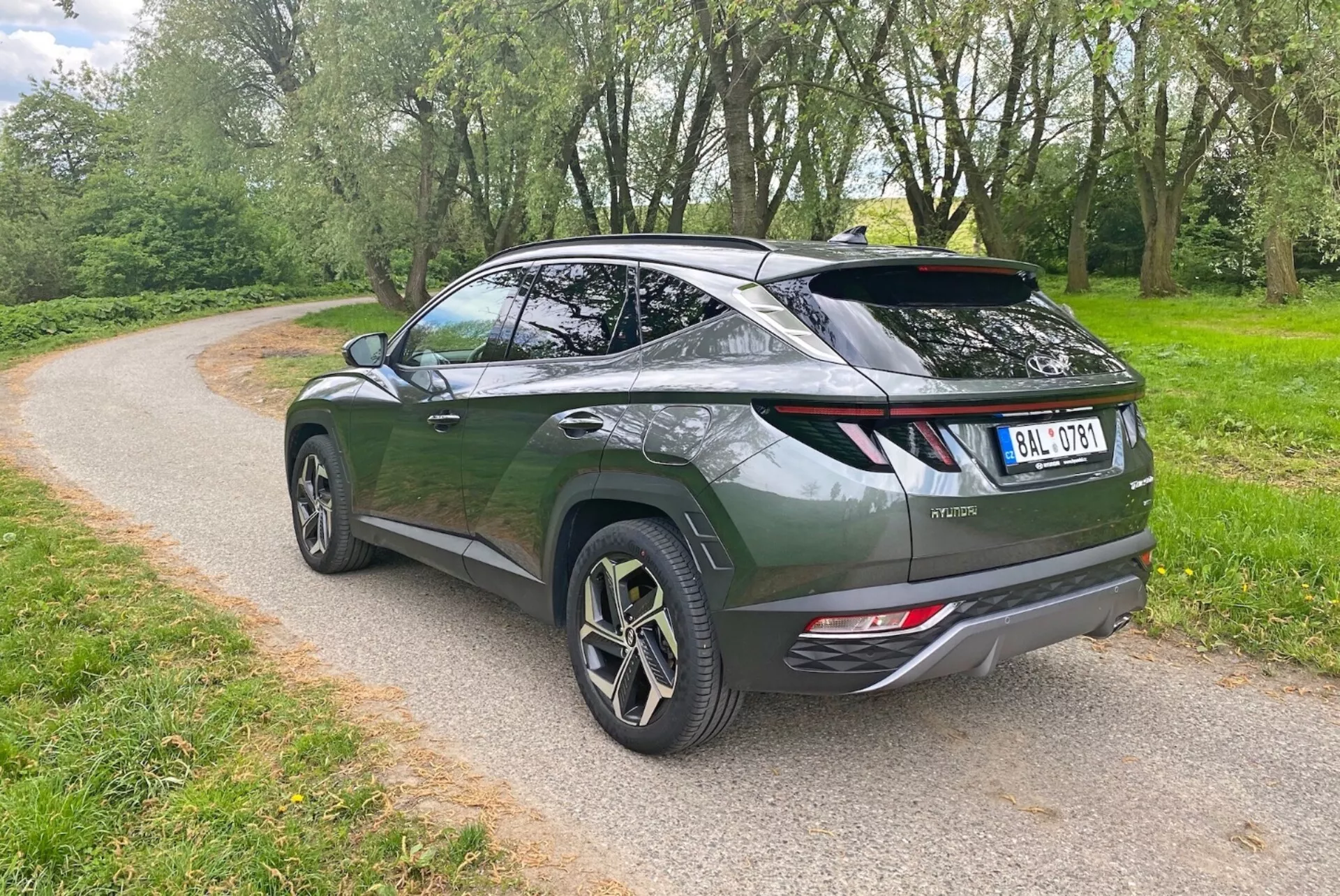 A képen egy modern crossover SUV látható háromnegyedes hátulnézetből, kiemelve az elegáns, aerodinamikus dizájnt és a lejtős tetővonalat. A sötétzöld jármű liftback-stílusú hátsó ajtóval, sportos spoilerral és többküllős kialakítású nagy könnyűfém kerekekkel rendelkezik. Látható elemek közé tartoznak a modern LED-es hátsó lámpák, a dupla kipufogócsövek és a hátsó ablaktörlő. Az SUV egy kavicsos úton parkol, amelyet zöld növényzet vesz körül, jelezve egy parkos vagy vidéki környezetet.