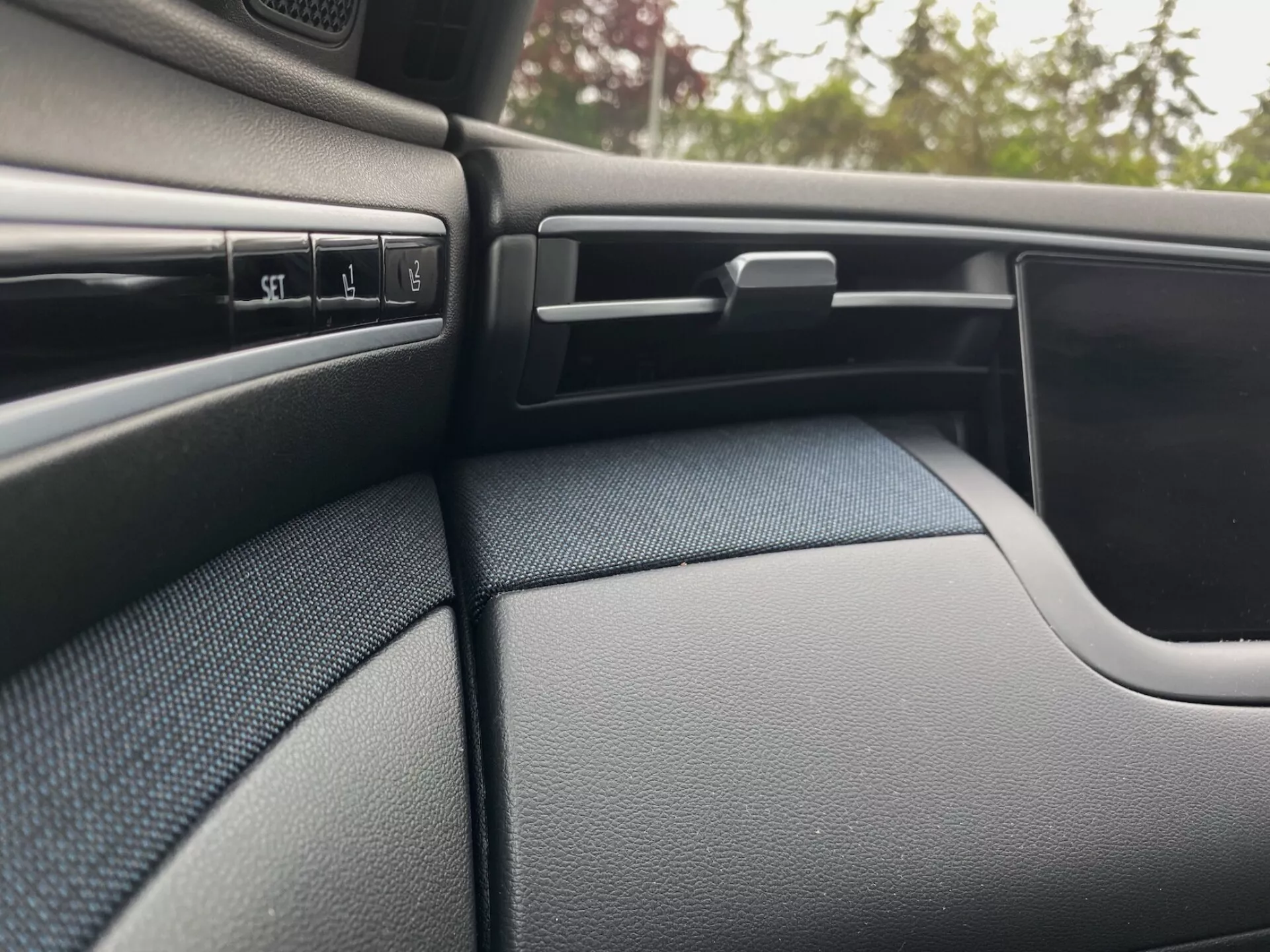 A Hyundai Tucson 2020 belső részlete, amely a sötét színű, texturált utasoldali műszerfalat és az ajtópanel egy részét mutatja. A jelenet kiemel több vezérlőgombot, egy állítható légbeömlőnyílást vízszintes lamellákkal, egy zárolható kesztyűtartót és az ajtópanel ívelt kialakítását, ami zökkenőmentesen illeszkedik a műszerfalhoz.