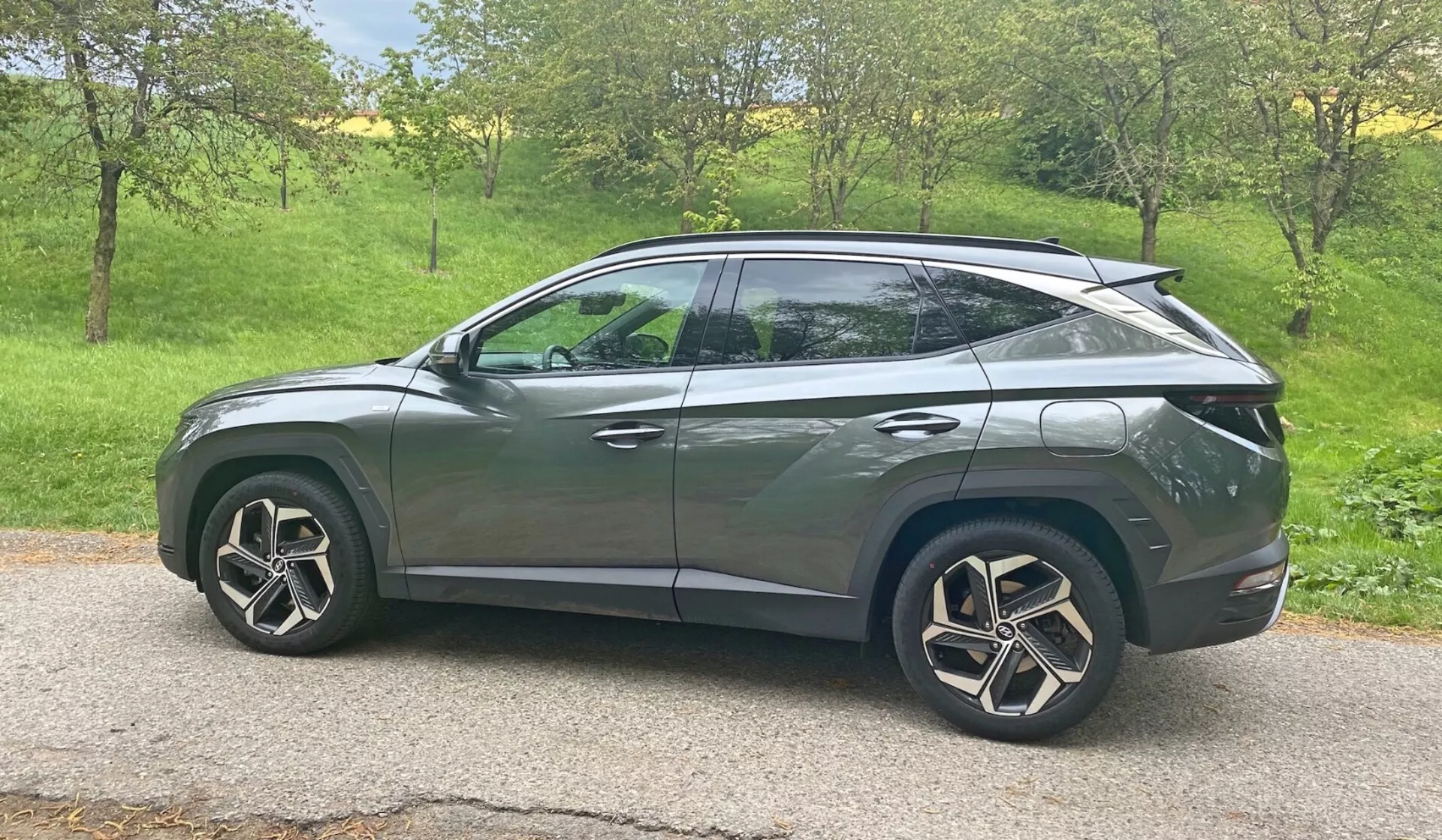 2020-as Hyundai Tucson 1.6 T-GDI Pure SUV oldalnézete természetes nappali fényben. A jármű egy füves területen helyezkedik el, fákkal a háttérben, ami vidéki vagy külvárosi környezetre utal. A modern dizájn jellemzői közé tartoznak a LED fényszórók, az oldalsó tükörbe integrált irányjelzők, a sötétített ablakok, az aerodinamikus könnyűfém keréktárcsák és a kupéhoz hasonló ferde tetővonal. Az elegáns karosszéria karaktervonalakkal és süllyesztett ajtókilincsekkel fejlett kulcs nélküli beléptetőrendszert sugall, míg a hátsó spoiler aerodinamikai fejlesztéseket jelez. Az autó központosan helyezkedik el a keretben, tiszta, akadálytalan kilátást nyújtva közepes magasságú perspektívából.